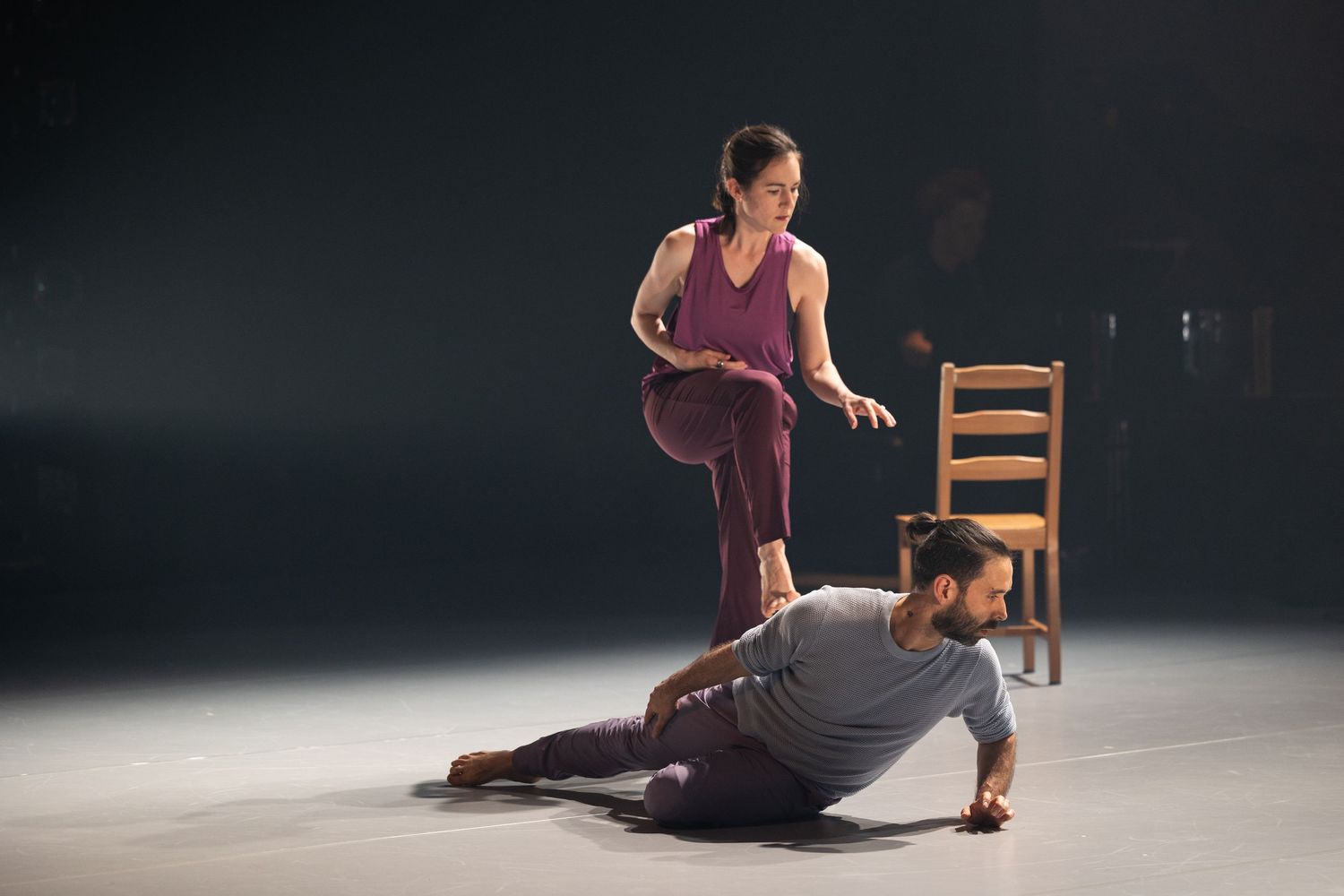 Two modern dancers perform a contemporary duet with a wooden chair against a dark background.