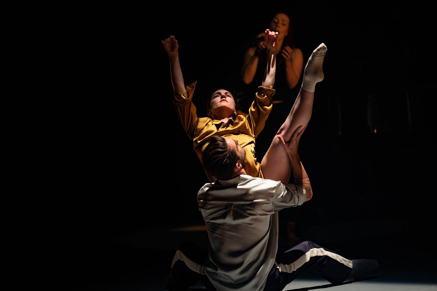 Dramatic dance performance with person reaching upward in yellow top against dark background.