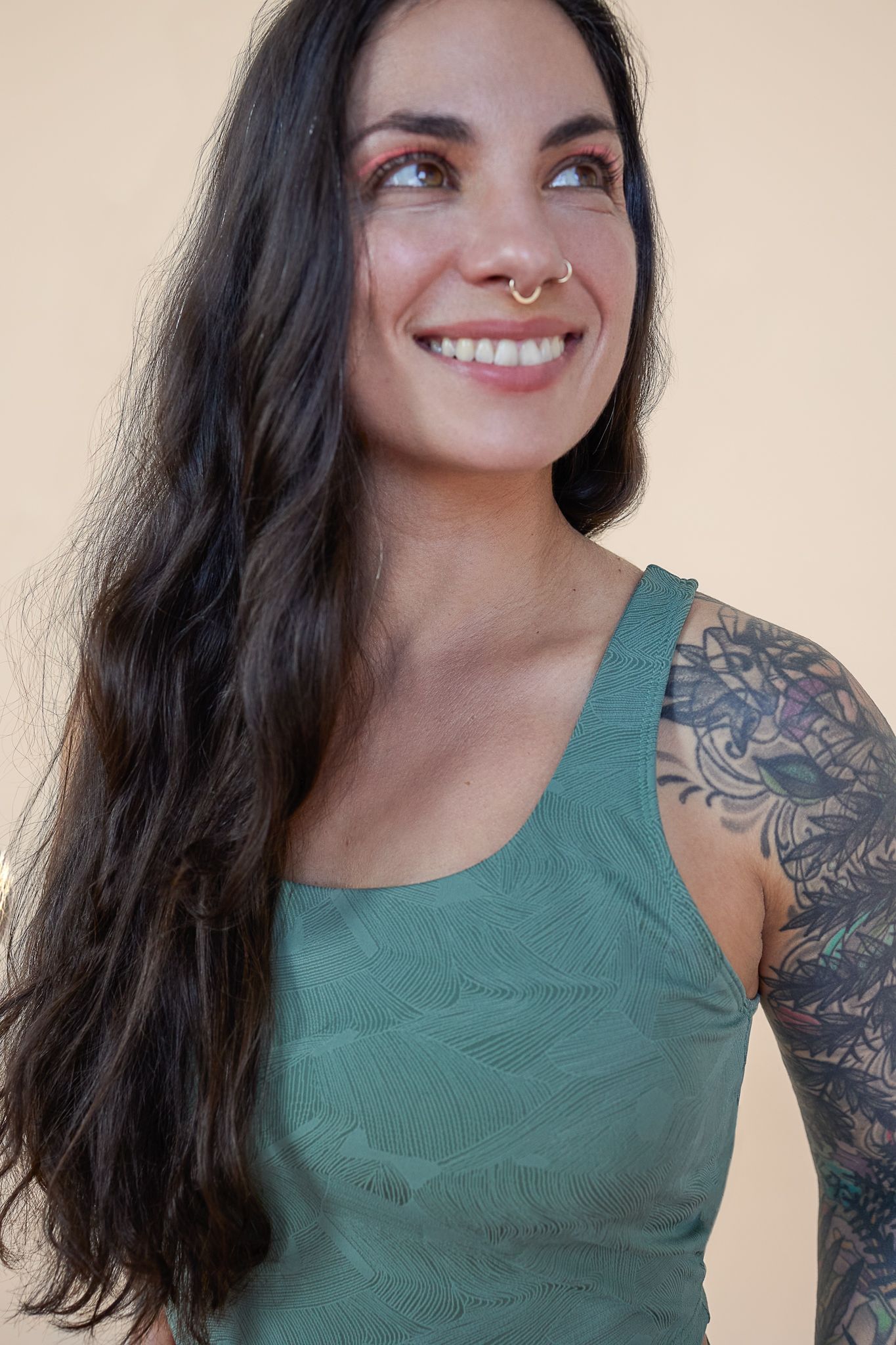Person with long dark hair wearing a green tank top smiling, showing nose piercing and colorful arm tattoo.