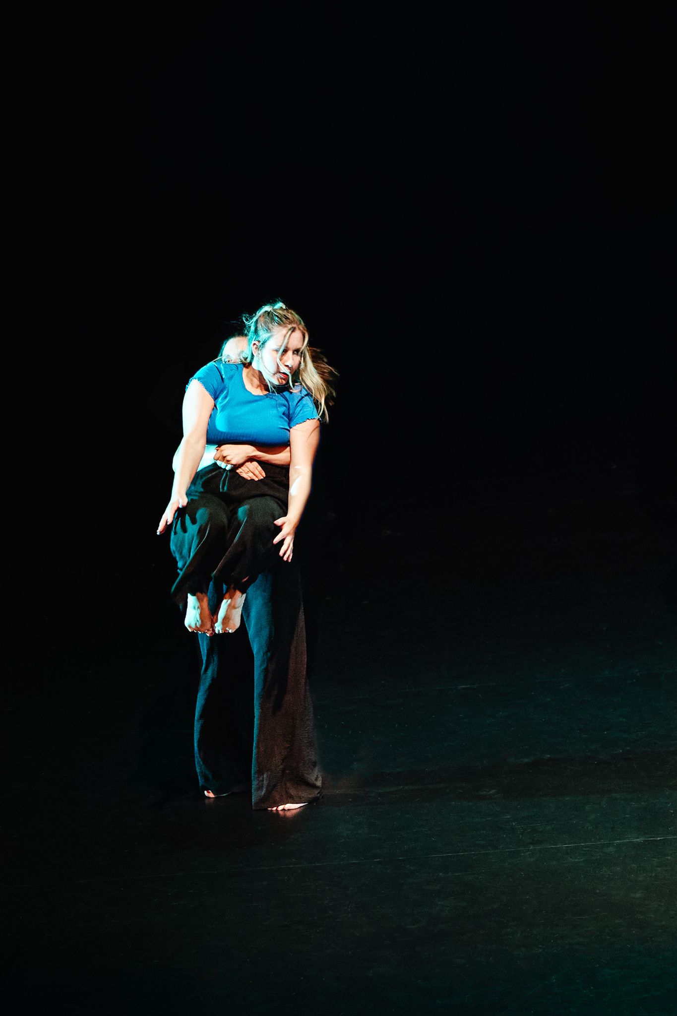 A dancer in turquoise and black performs on a dark stage, bending forward in an expressive contemporary dance pose.