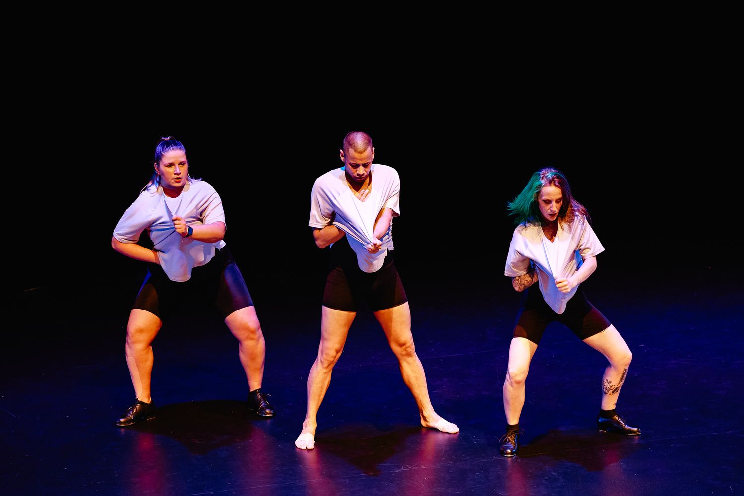Three performers in white shirts and black shorts strike synchronized dance poses on a dark stage.