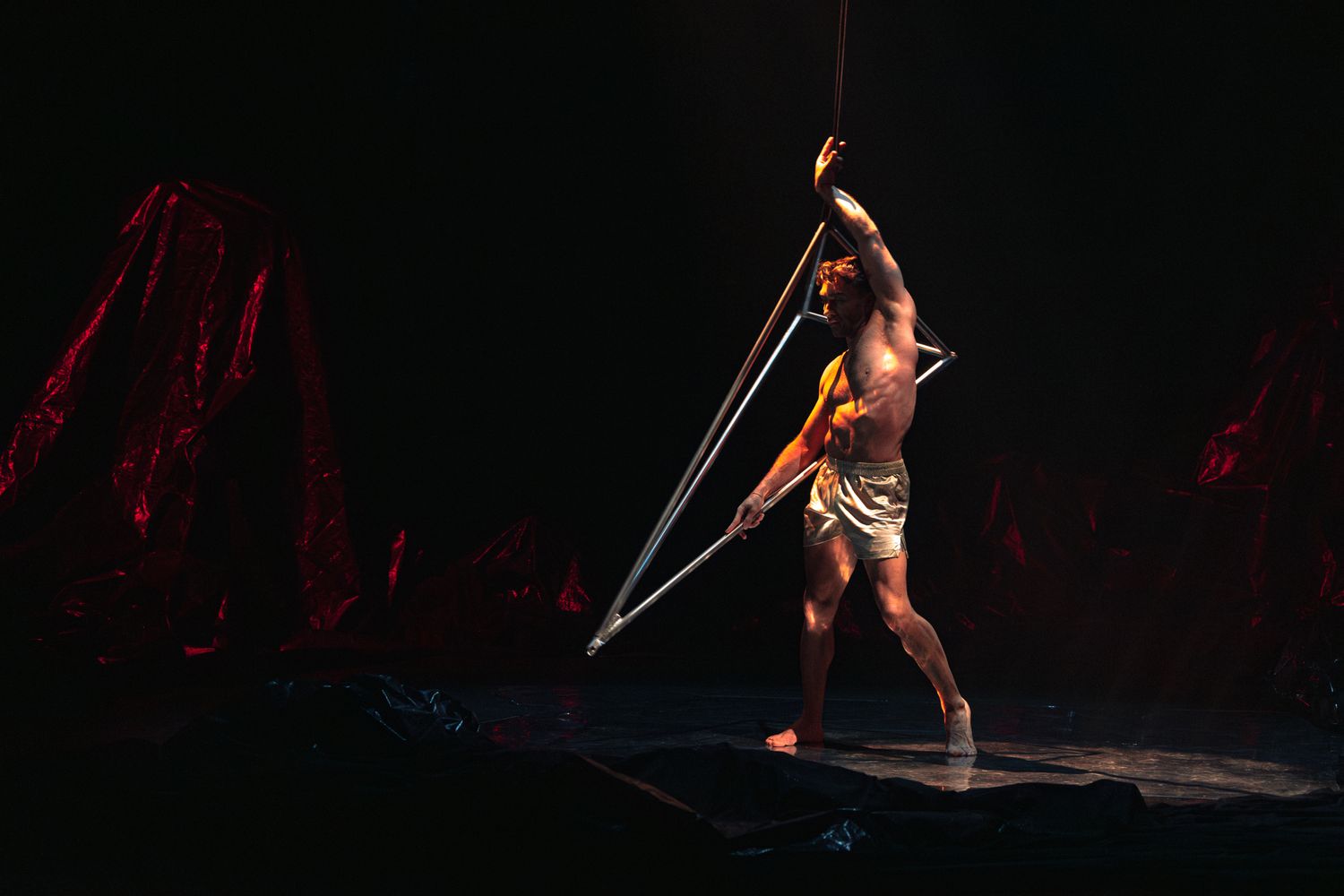 Muscular circus performer in gold costume balances gracefully on aerial straps against dark dramatic stage lighting.