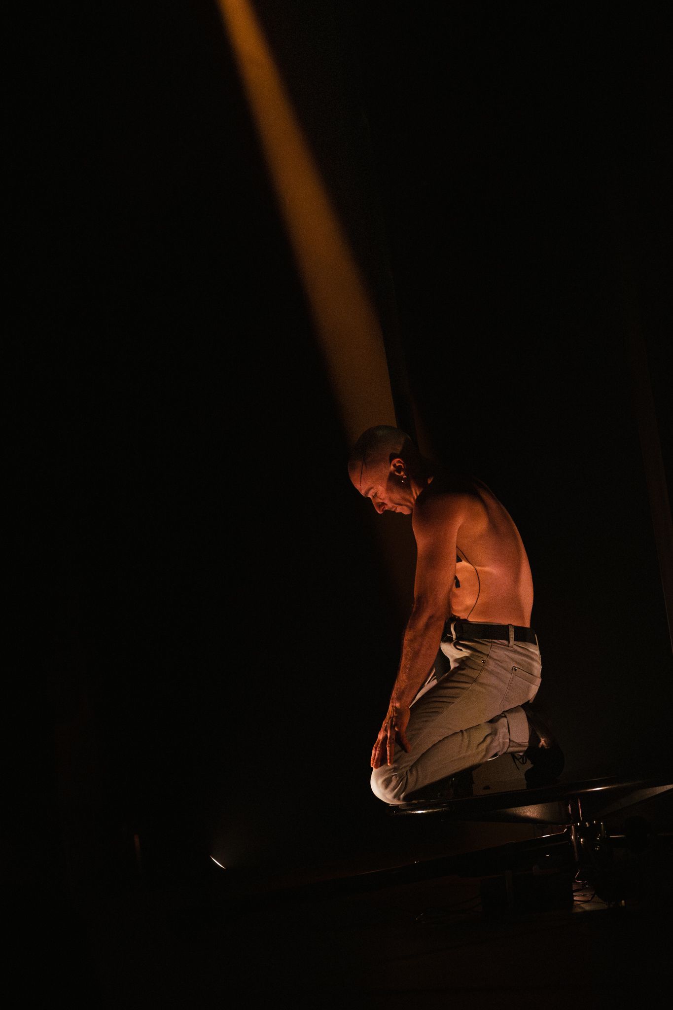 Performer shirtless in dramatic lighting crouches on stage during theatrical performance against dark background.