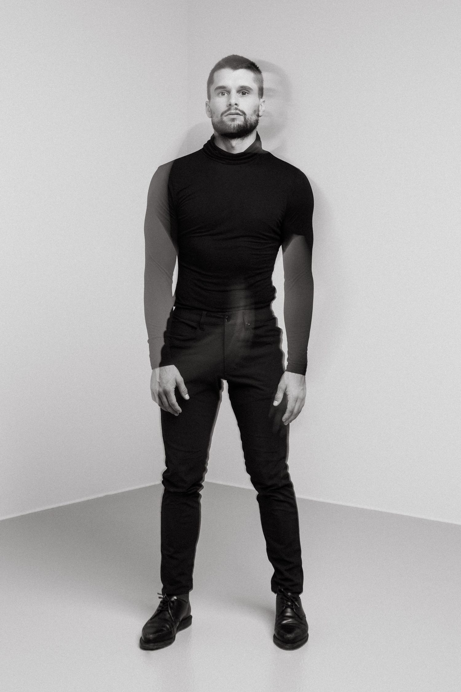 Black and white studio portrait of person wearing all black turtleneck and pants standing against light background.