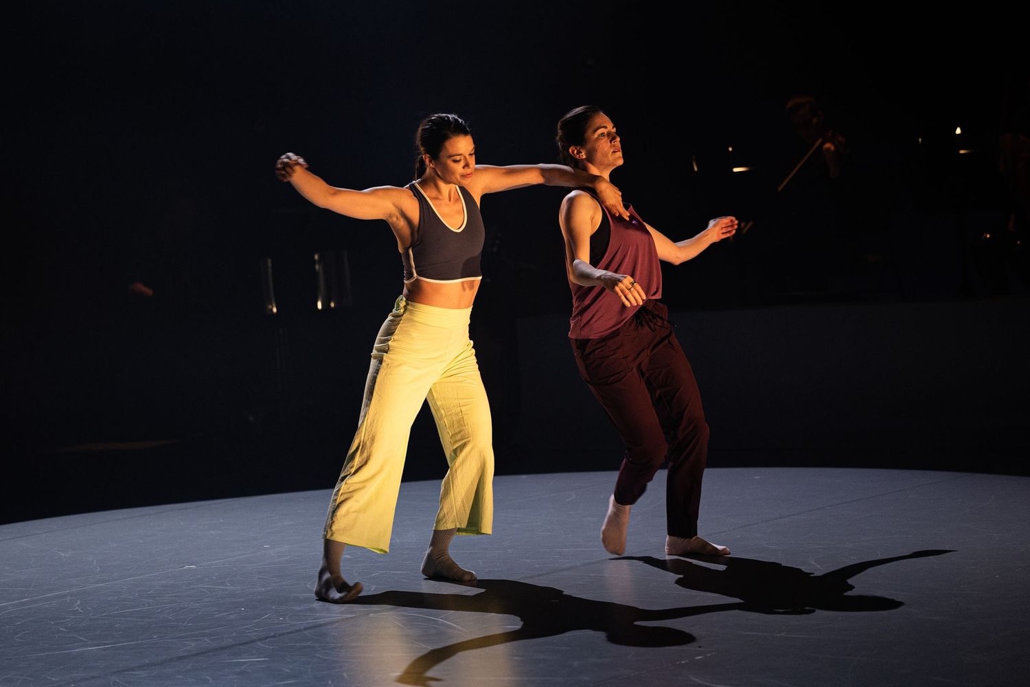 Two dancers perform in spotlight on dark stage wearing yellow and burgundy outfits while striking synchronized poses.