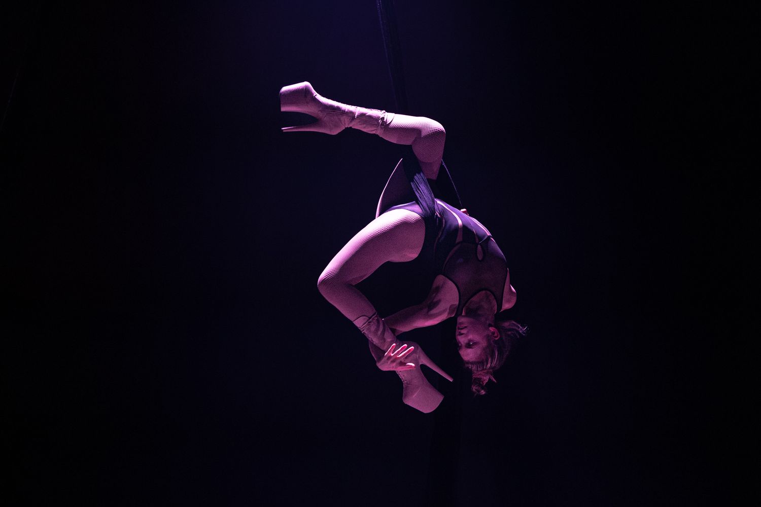A performer in purple lighting executes a graceful aerial acrobatic maneuver against a dark background.