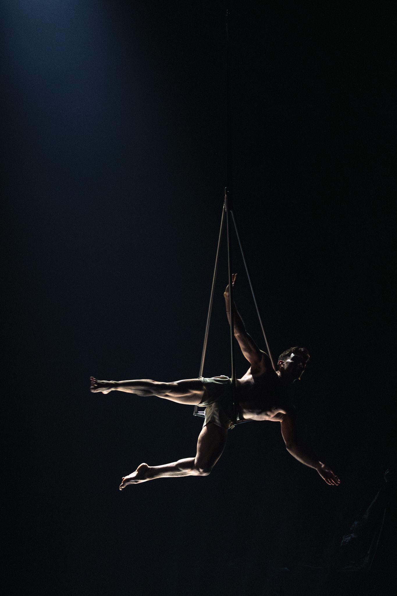 An aerial performer suspended by ropes gracefully poses against a dark backdrop in a dramatic circus performance.