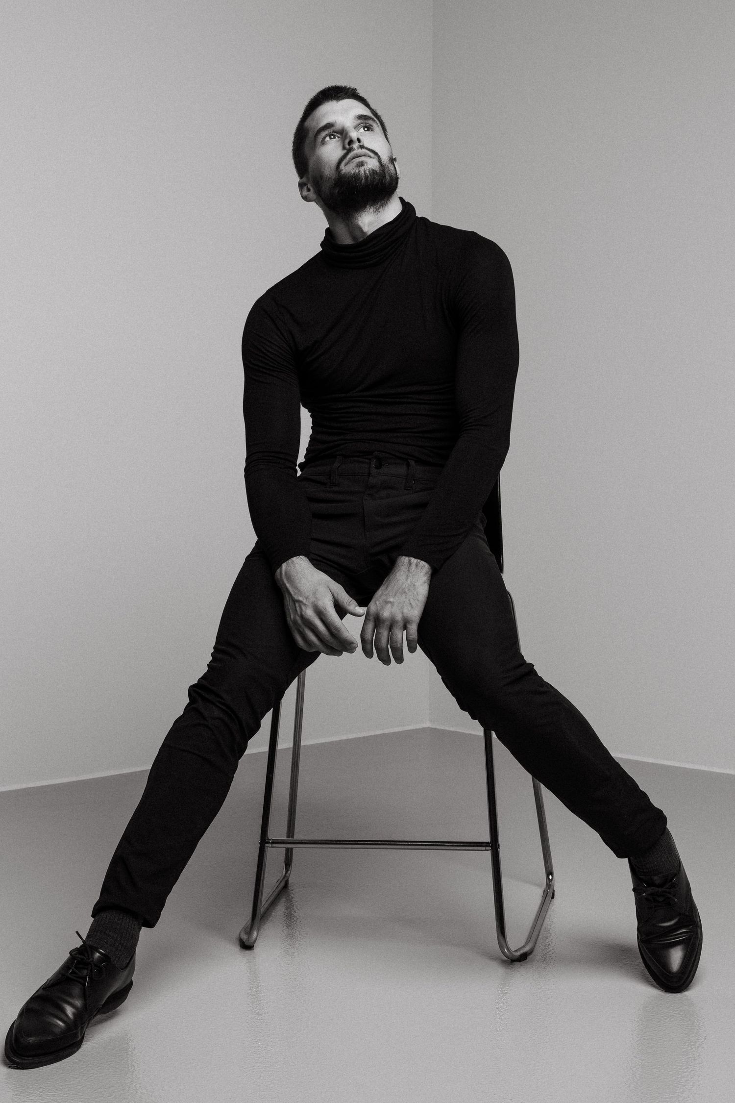 Dramatic black and white portrait of person in all black attire seated on stool against minimal studio background.