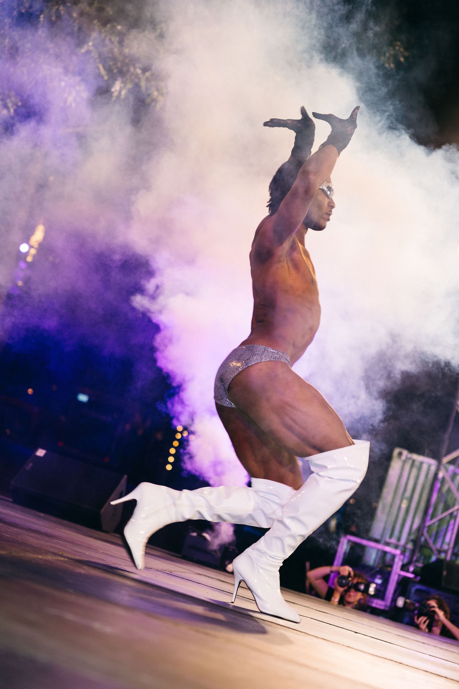 A performer in white boots dances energetically onstage amidst dramatic purple smoke effects and stage lighting.
