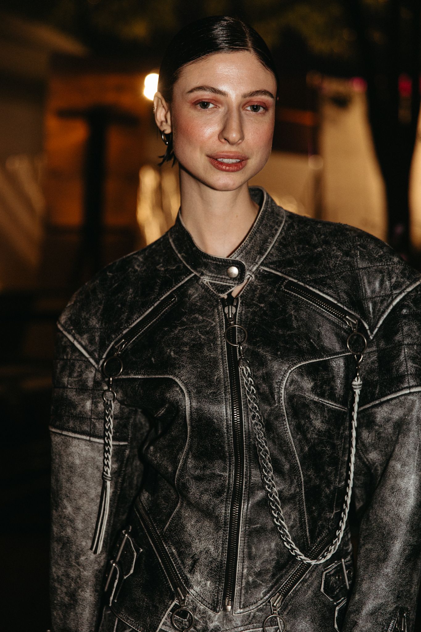 A figure wearing a distressed black leather jacket with intricate stitching and metallic details poses in warm lighting.