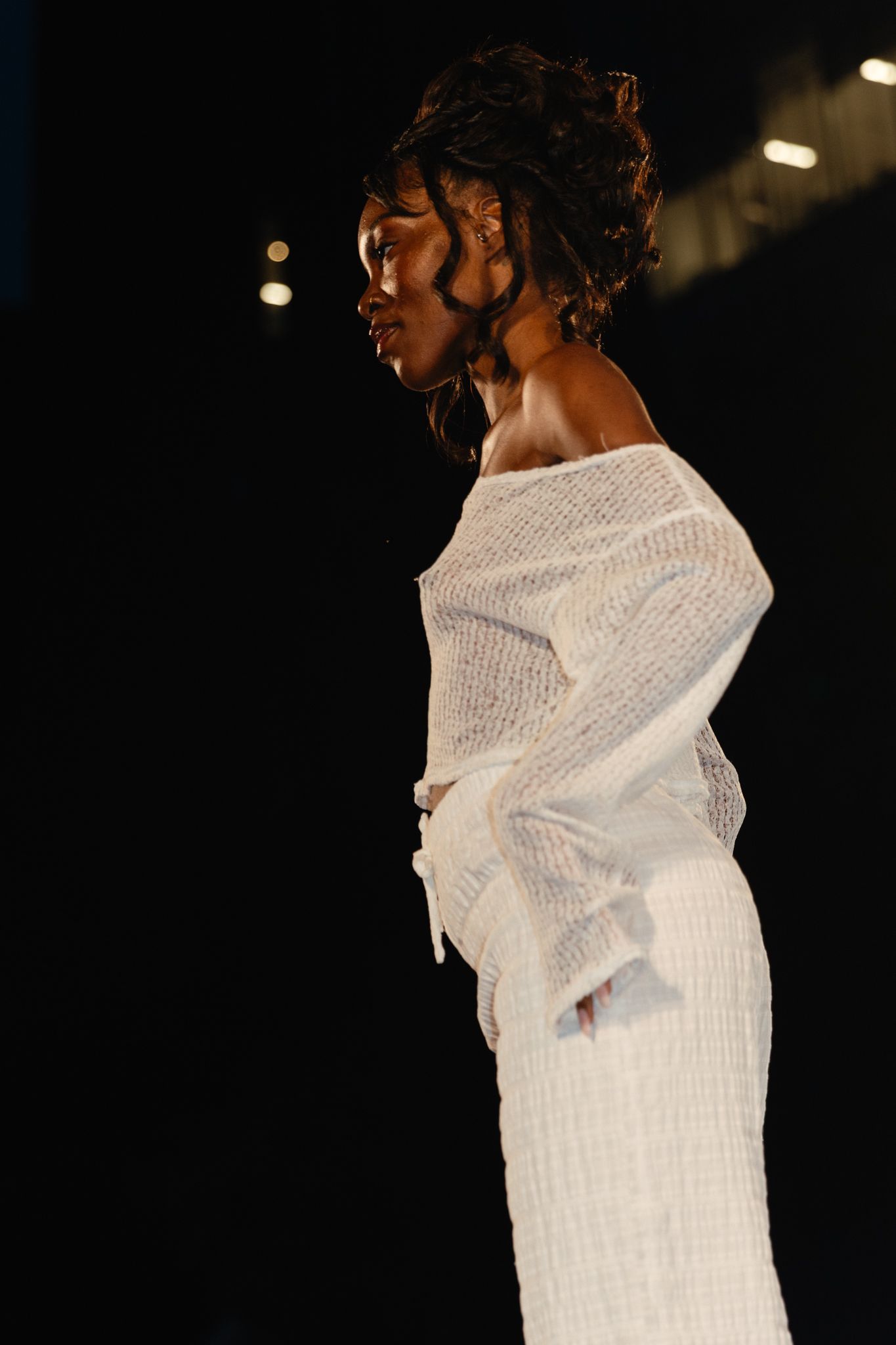 Side profile of a model in an elegant white knit dress on a dark stage during a fashion show.
