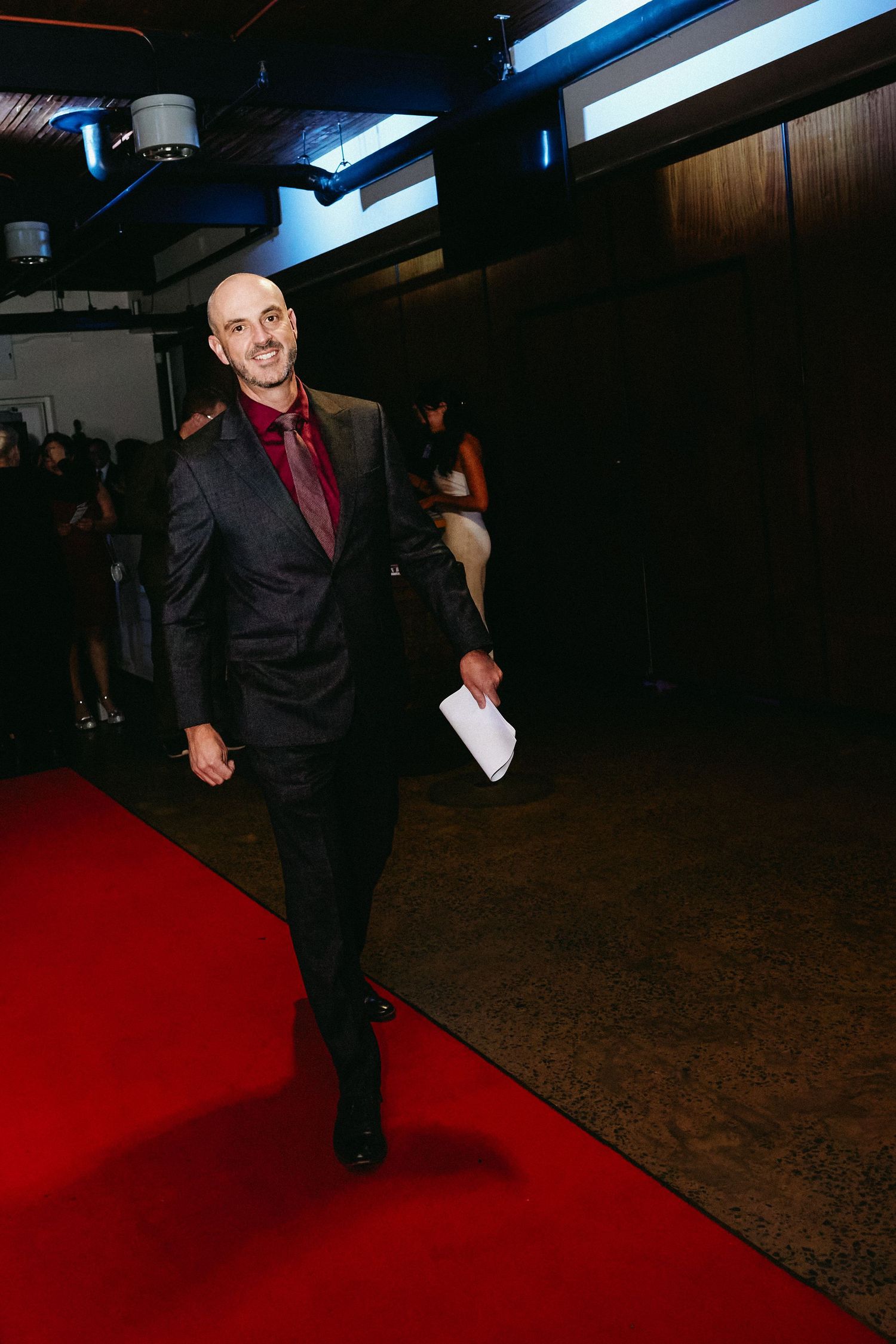 A person dressed in black attire performs energetic dance moves on a red carpet at a dark event venue.