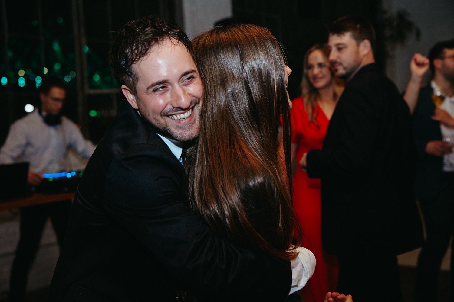 Two people dancing and embracing at a nighttime social event with party guests visible in the background.
