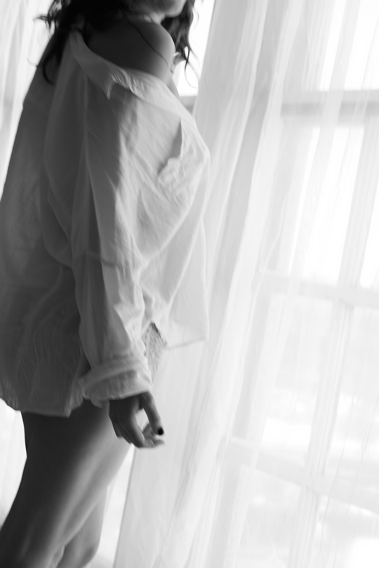 A couple in white clothing embraces next to sheer curtains in a tender black and white moment.