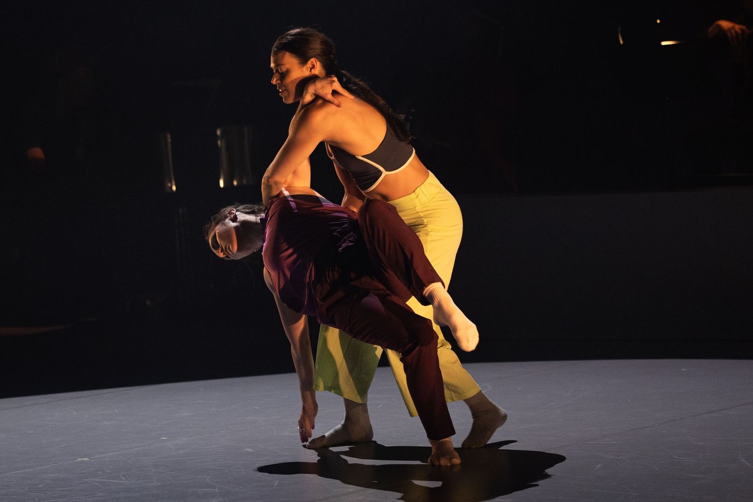 Dancers in yellow and brown costumes perform a contemporary duet on a dark stage.