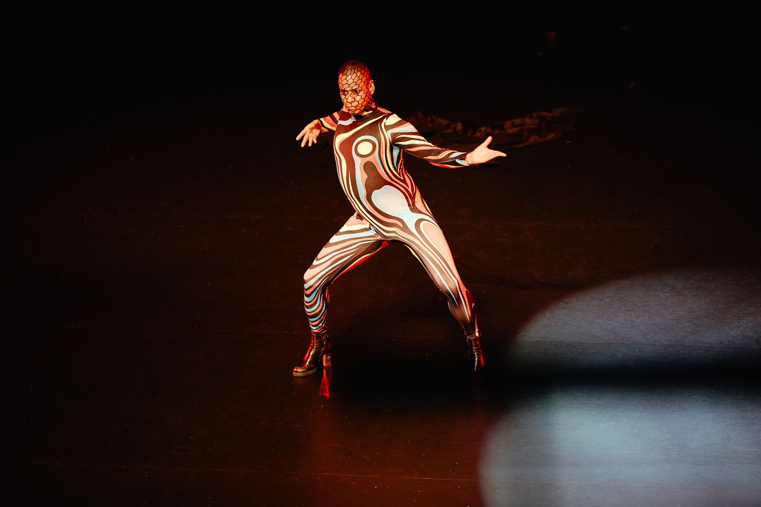 A dancer in a futuristic white costume performs an expressive movement on a dimly lit stage.