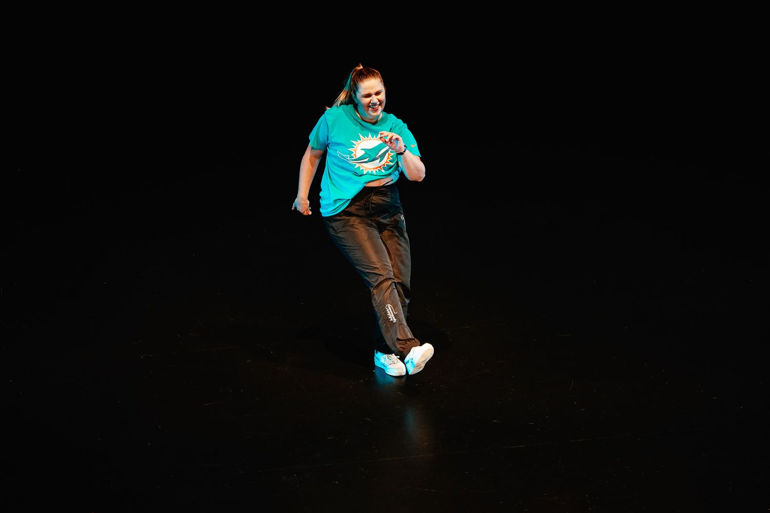 A person wearing a teal t-shirt and brown pants appears to be jumping or dancing against a black background.