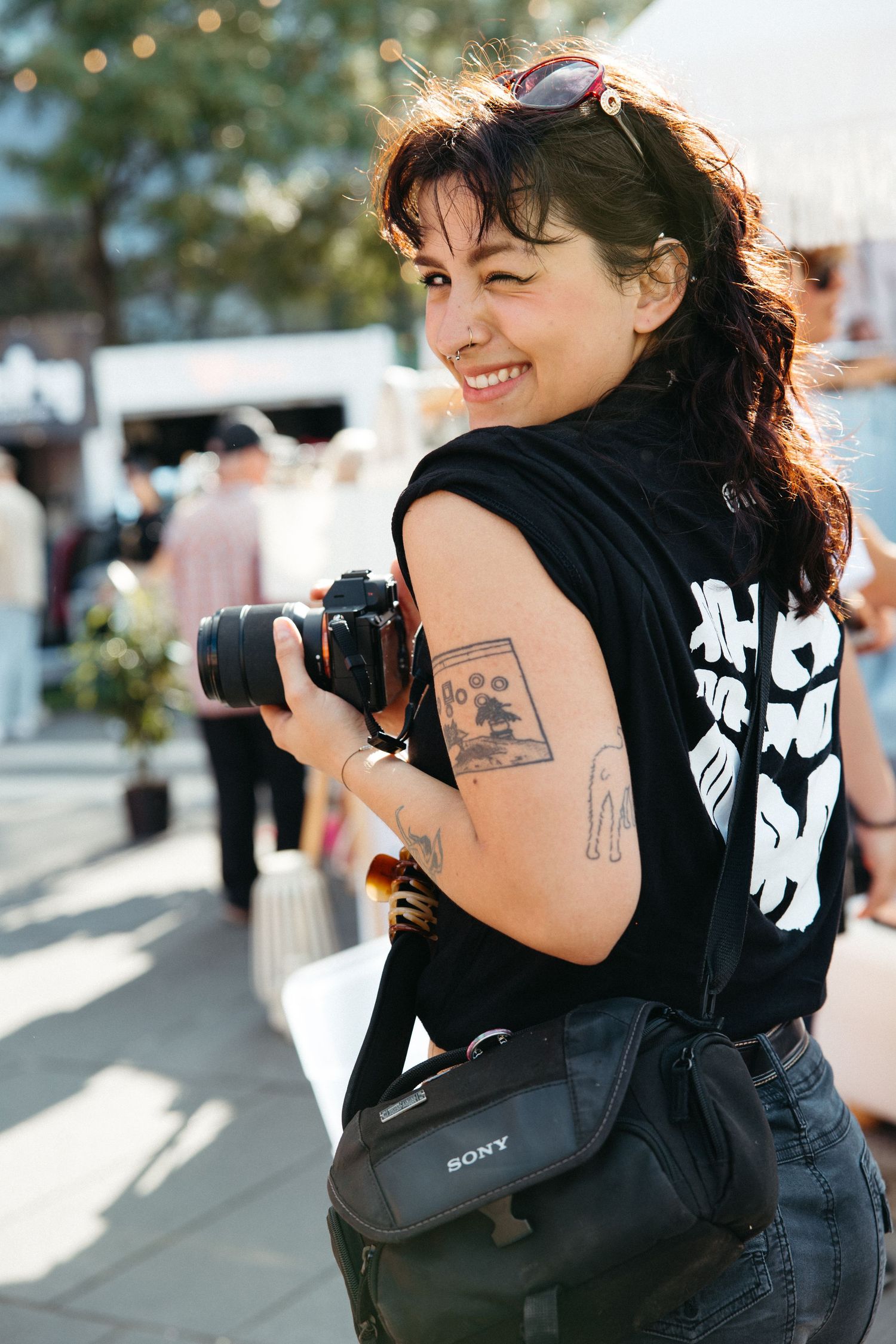 A photographer with dark curly hair and tattoos smiles while holding a camera outdoors on a sunny day.