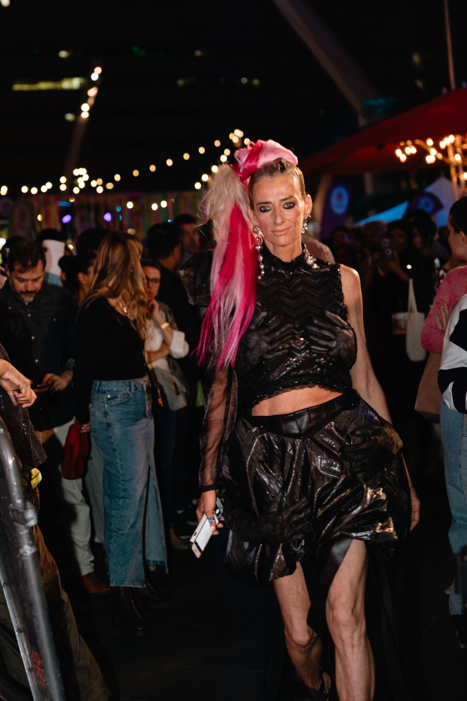 A person in a black outfit with pink hair dances at a nighttime event with string lights overhead.