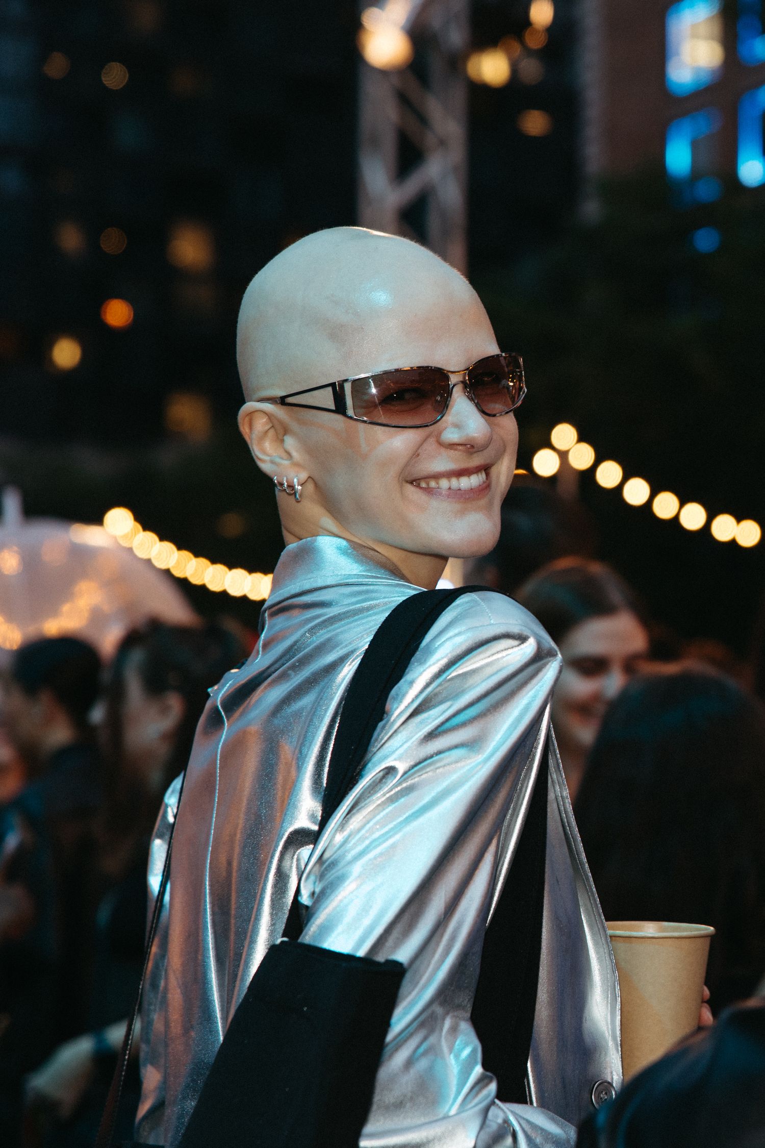 Person in metallic silver jacket and sunglasses smiles at nighttime event with glowing lights in background.