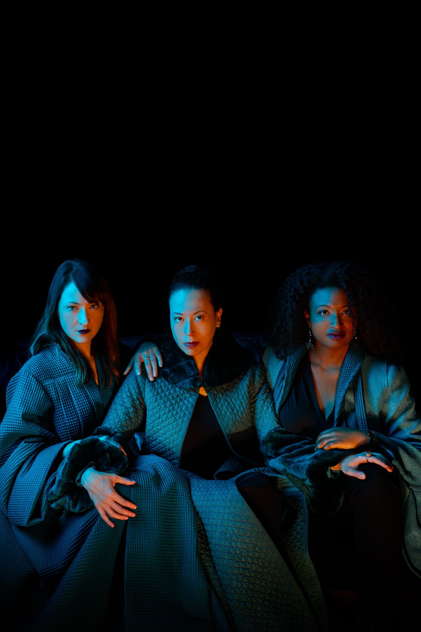 Dramatic portrait of three figures seated together under moody blue lighting against a dark background.
