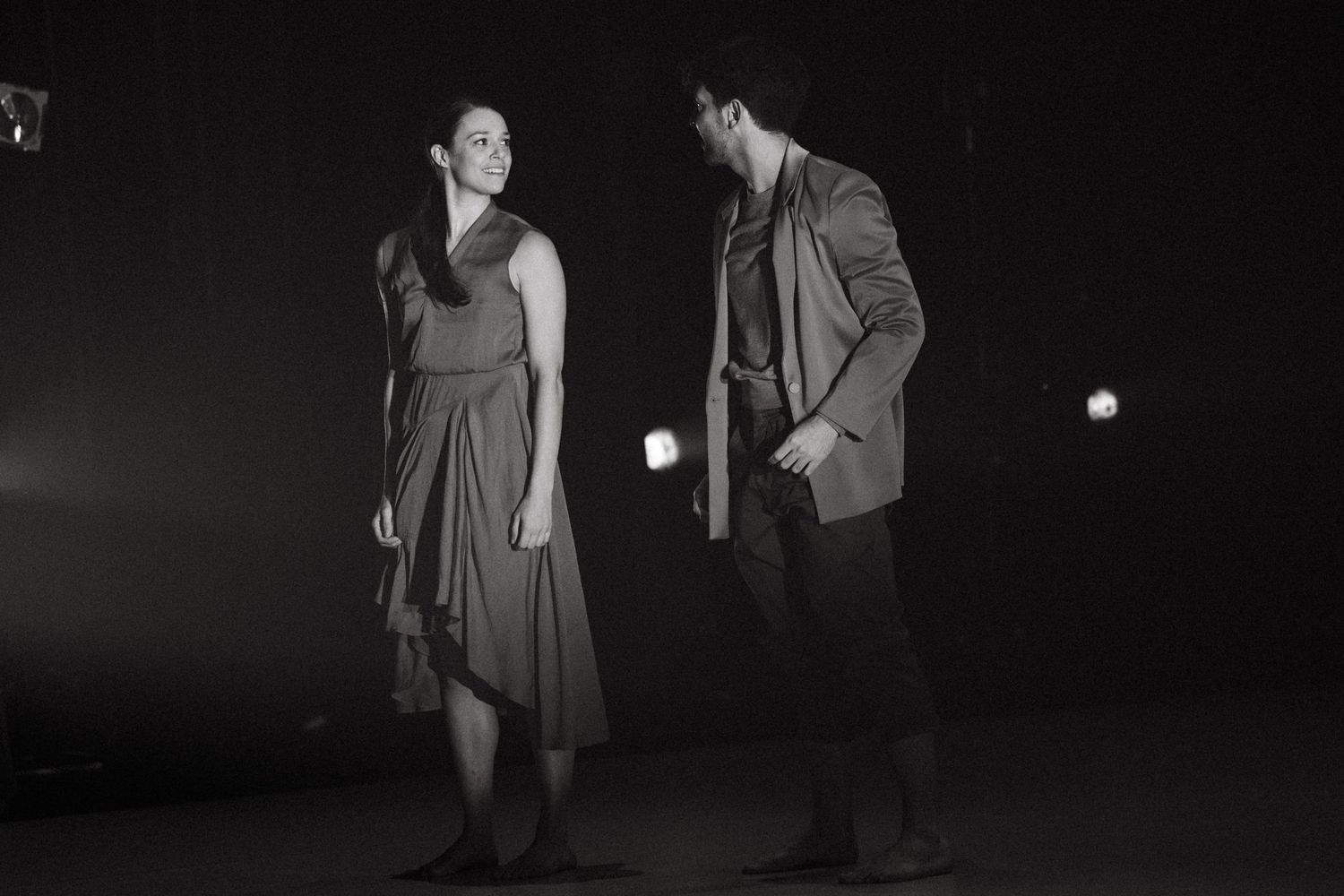 Two performers stand in dramatic lighting on a dark stage in what appears to be a theatrical scene or performance.