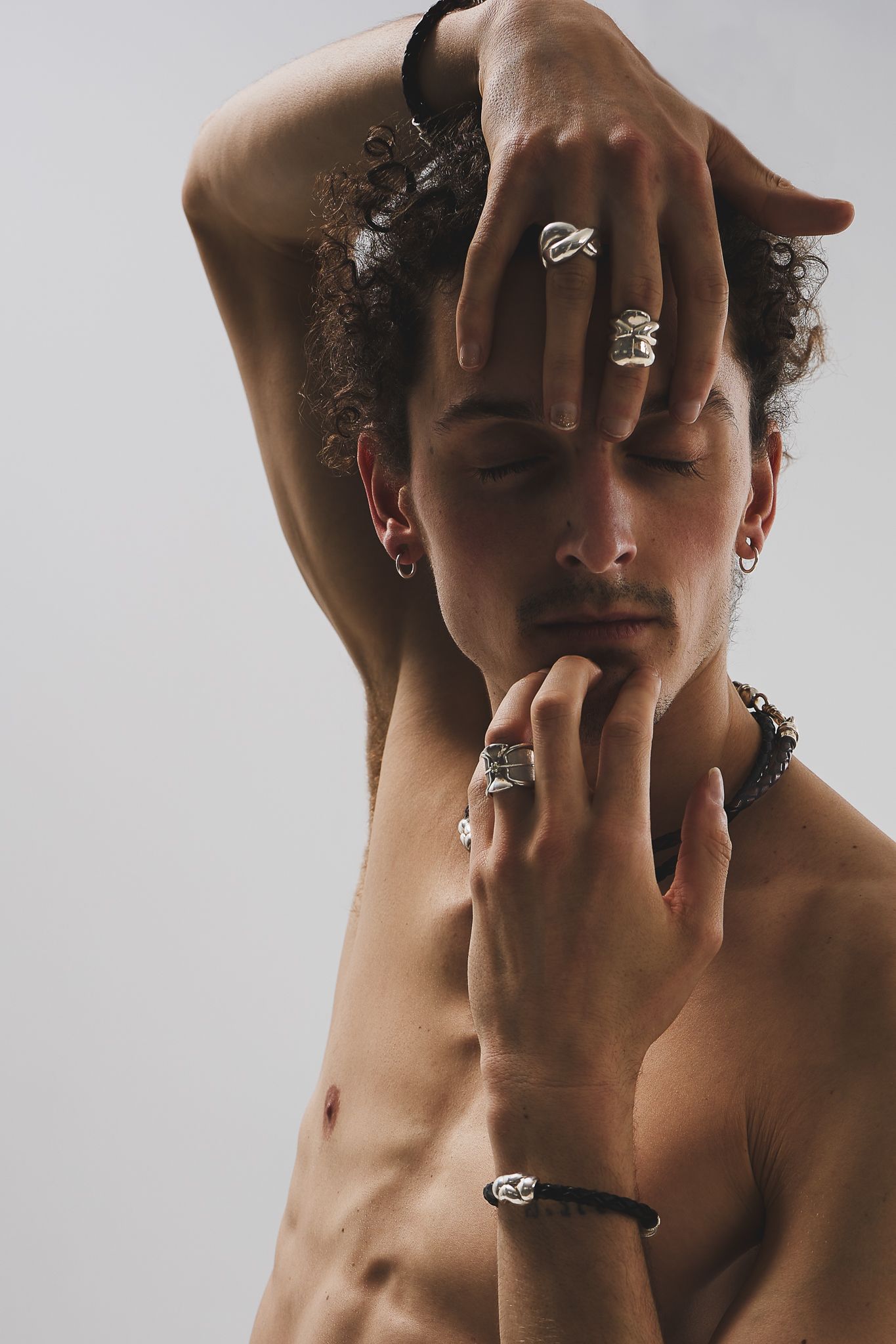 Artistic portrait showing jewelry and accessories on hands against bare torso in moody lighting and gray background.