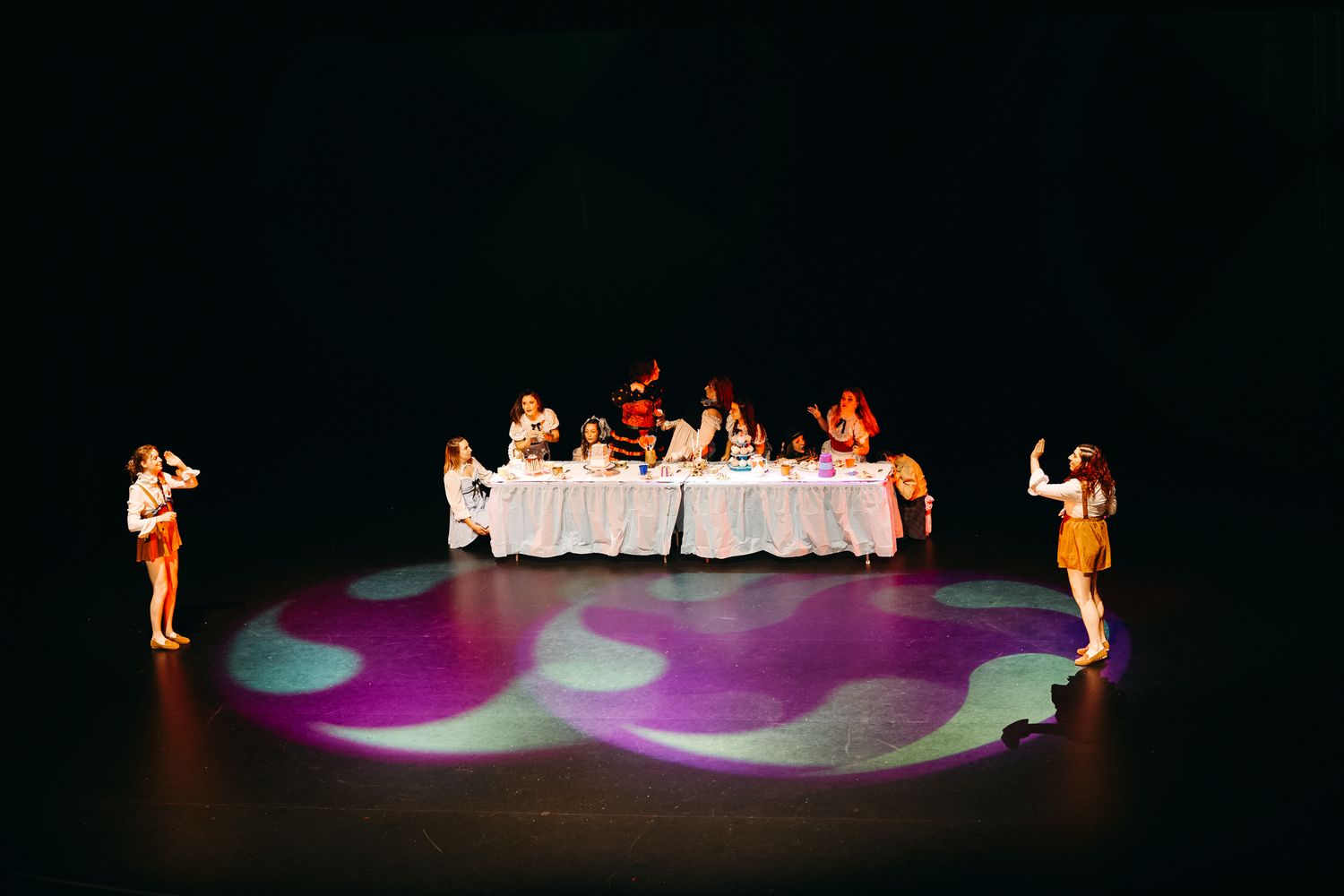 Theatrical performance showing dining scene at long table with performers in spotlight on dark stage with purple lighting effects.