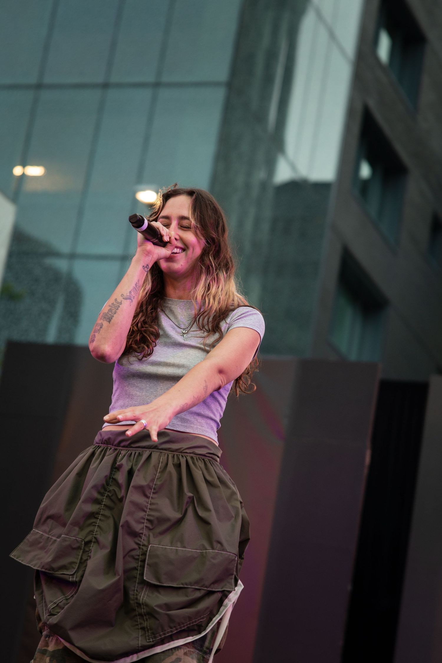 Singer performs on stage wearing a grey t-shirt and cargo skirt while holding a microphone during an outdoor concert.