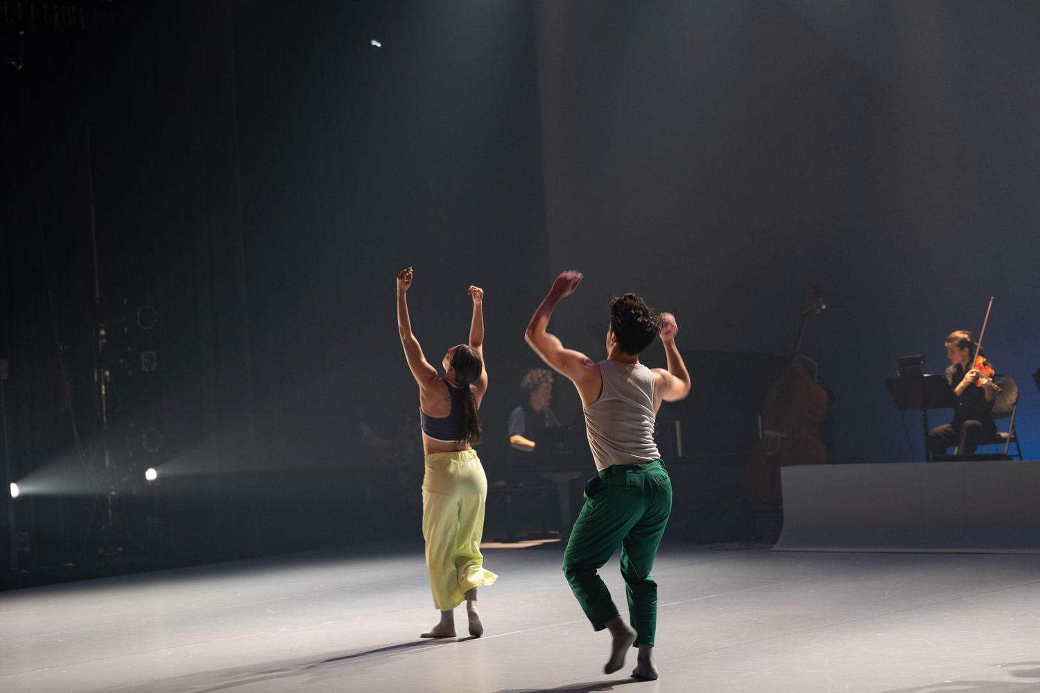 Two dancers perform an energetic routine on a dark stage with dramatic lighting and silhouetted figures in the background.