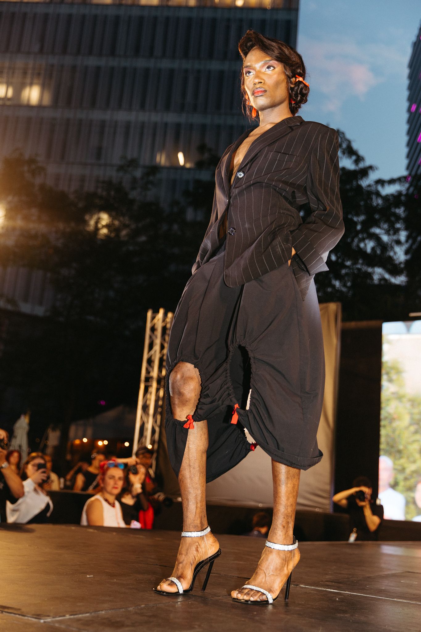 Model walks runway in sleek black dress and stilettos during evening fashion show with city buildings in background.