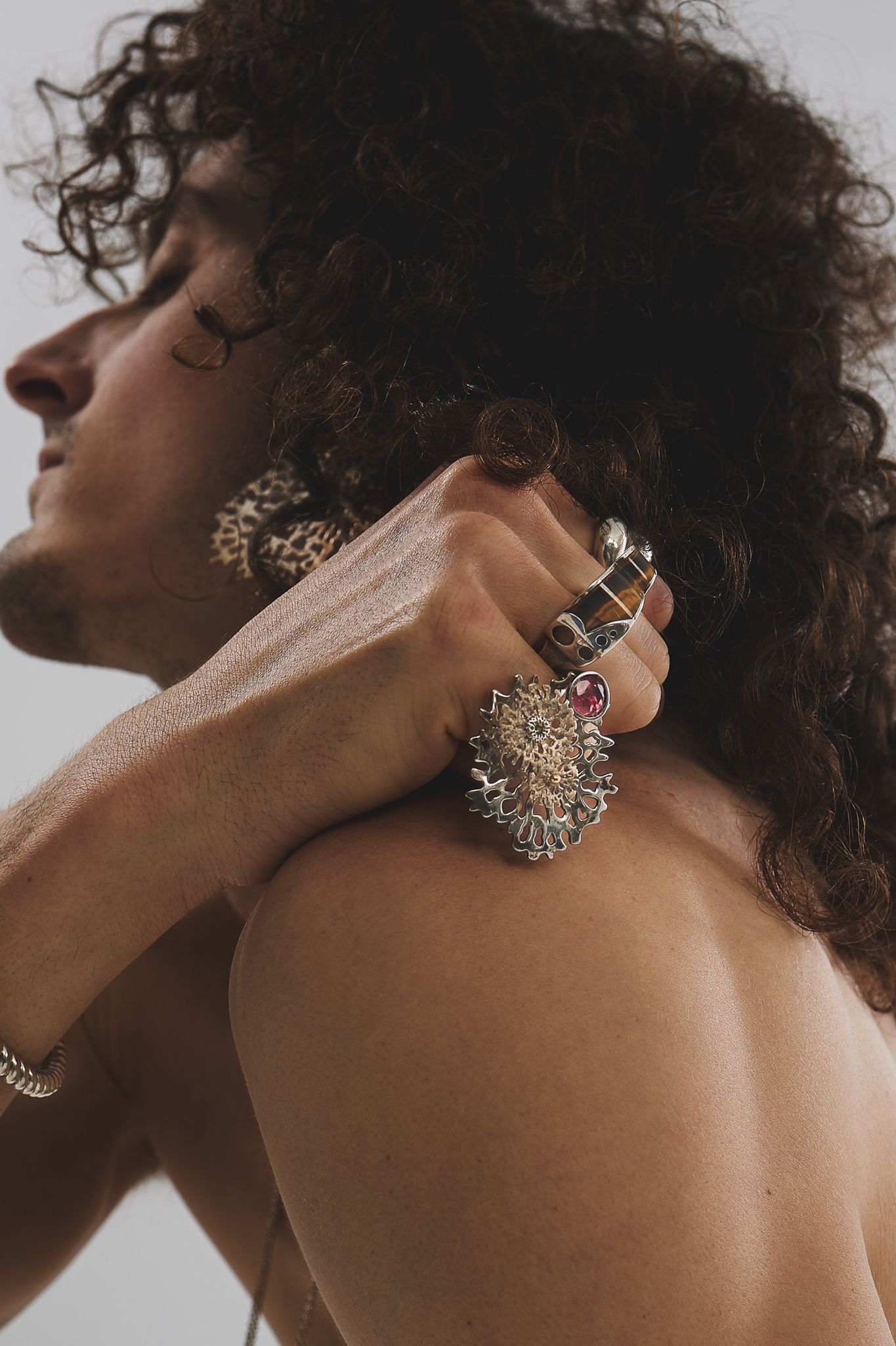 Close-up artistic fashion photograph showcasing ornate statement jewelry and curly hair against a light background.
