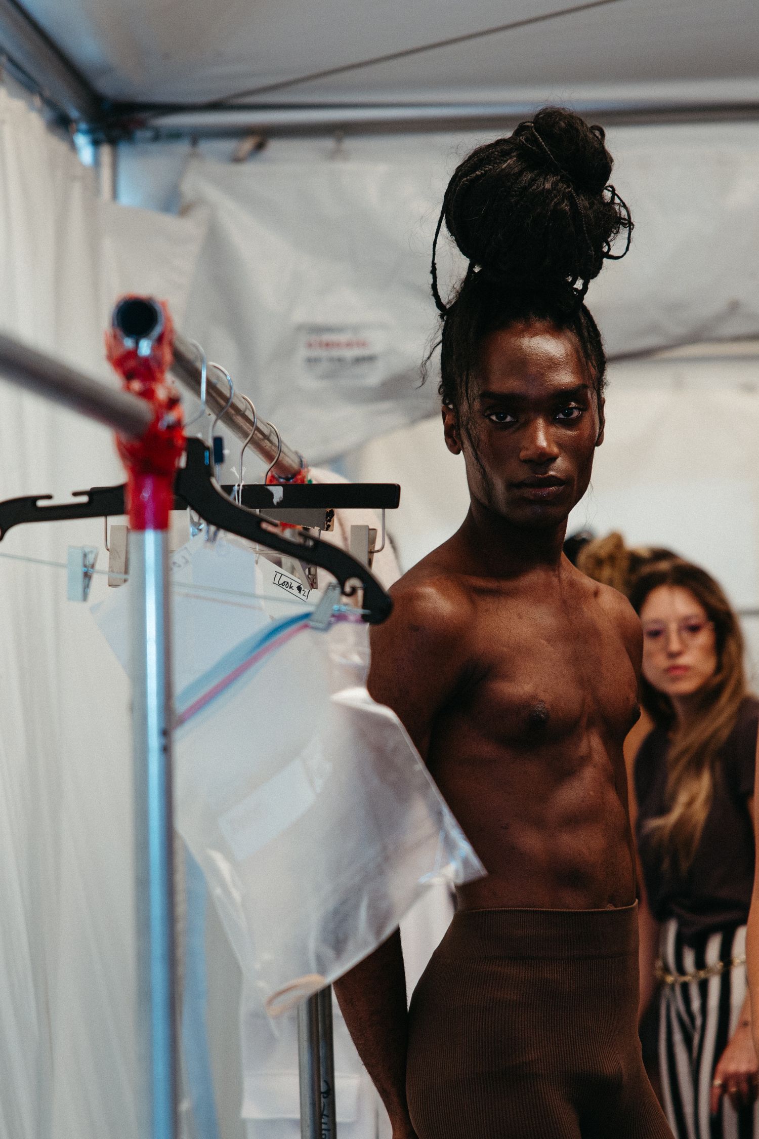 Backstage at a fashion show with a model standing near garment racks and seamstress tools.