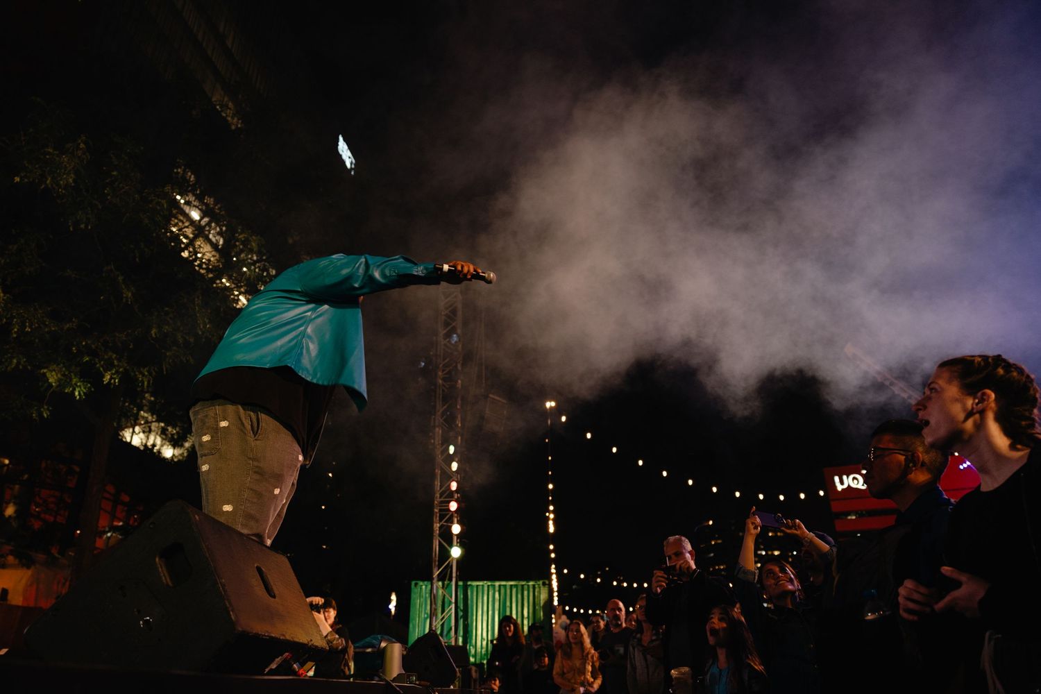 Concert crowd silhouetted against moody atmospheric lighting and smoke with string lights overhead during nighttime performance.