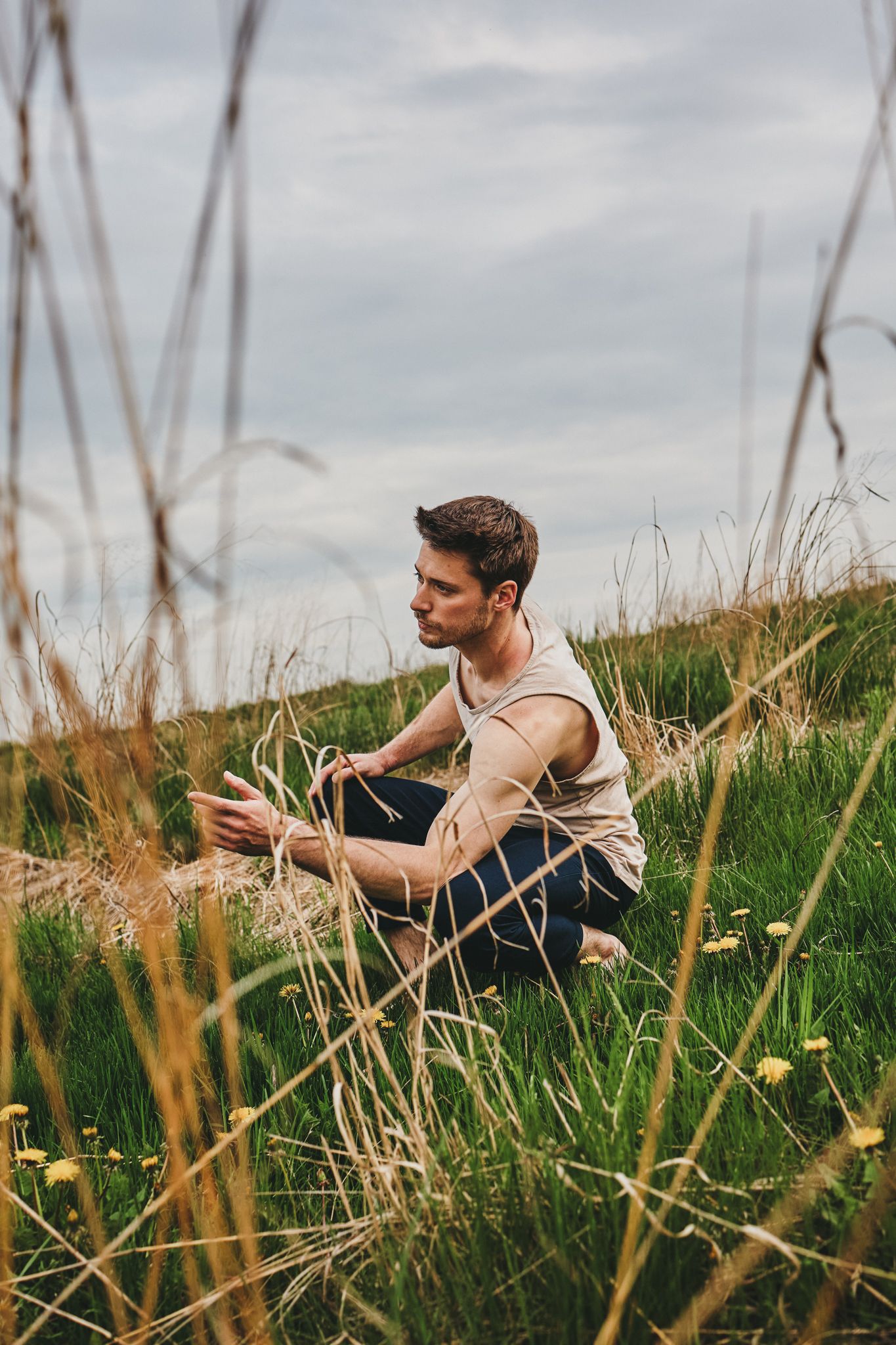 Person in a white tank top lying in a grassy meadow with tall grass and cloudy skies in the background.