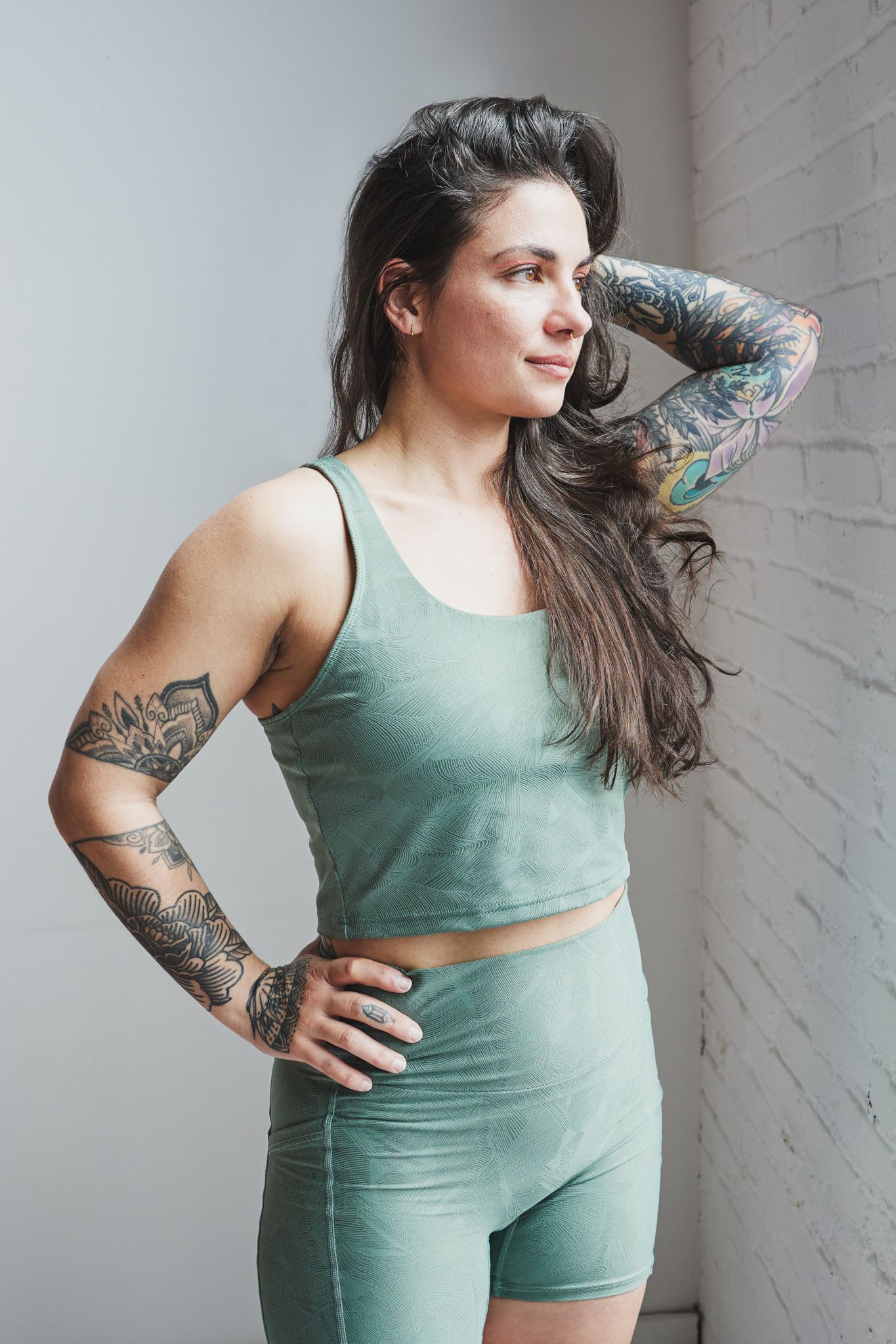 Athletic person in matching sage activewear set shows colorful sleeve tattoos while posing by white brick wall.