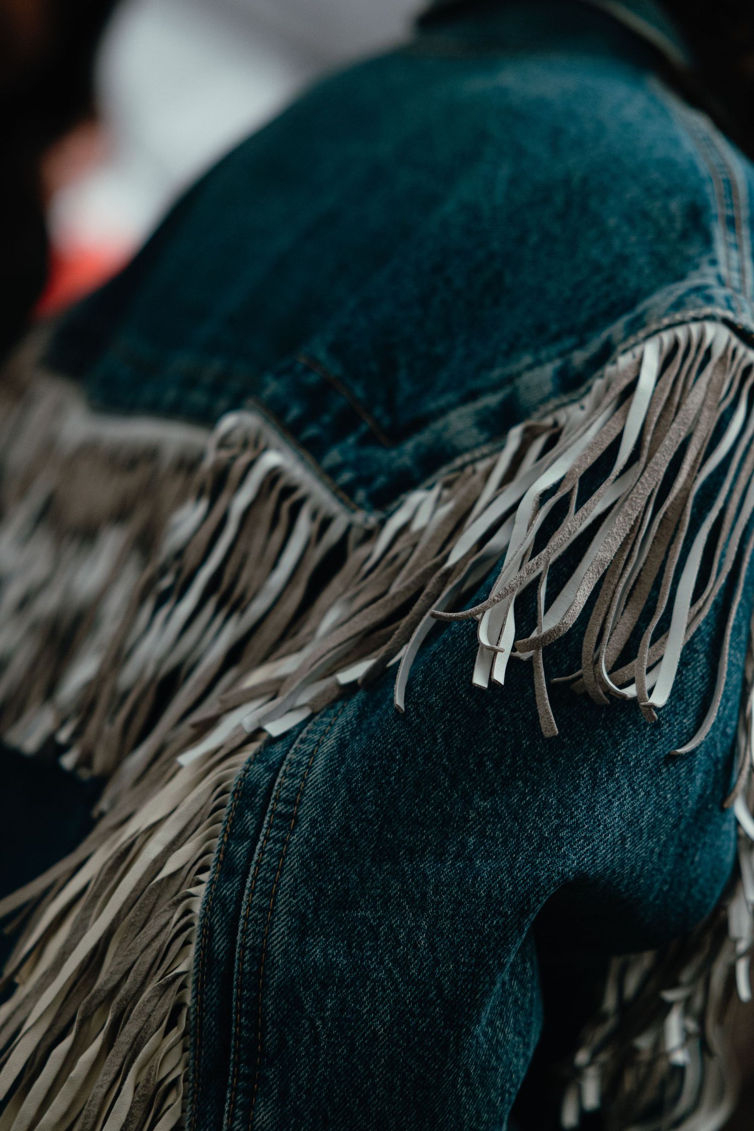 Close-up detail of a western jacket with teal fabric and beige leather fringe trim in dramatic lighting.