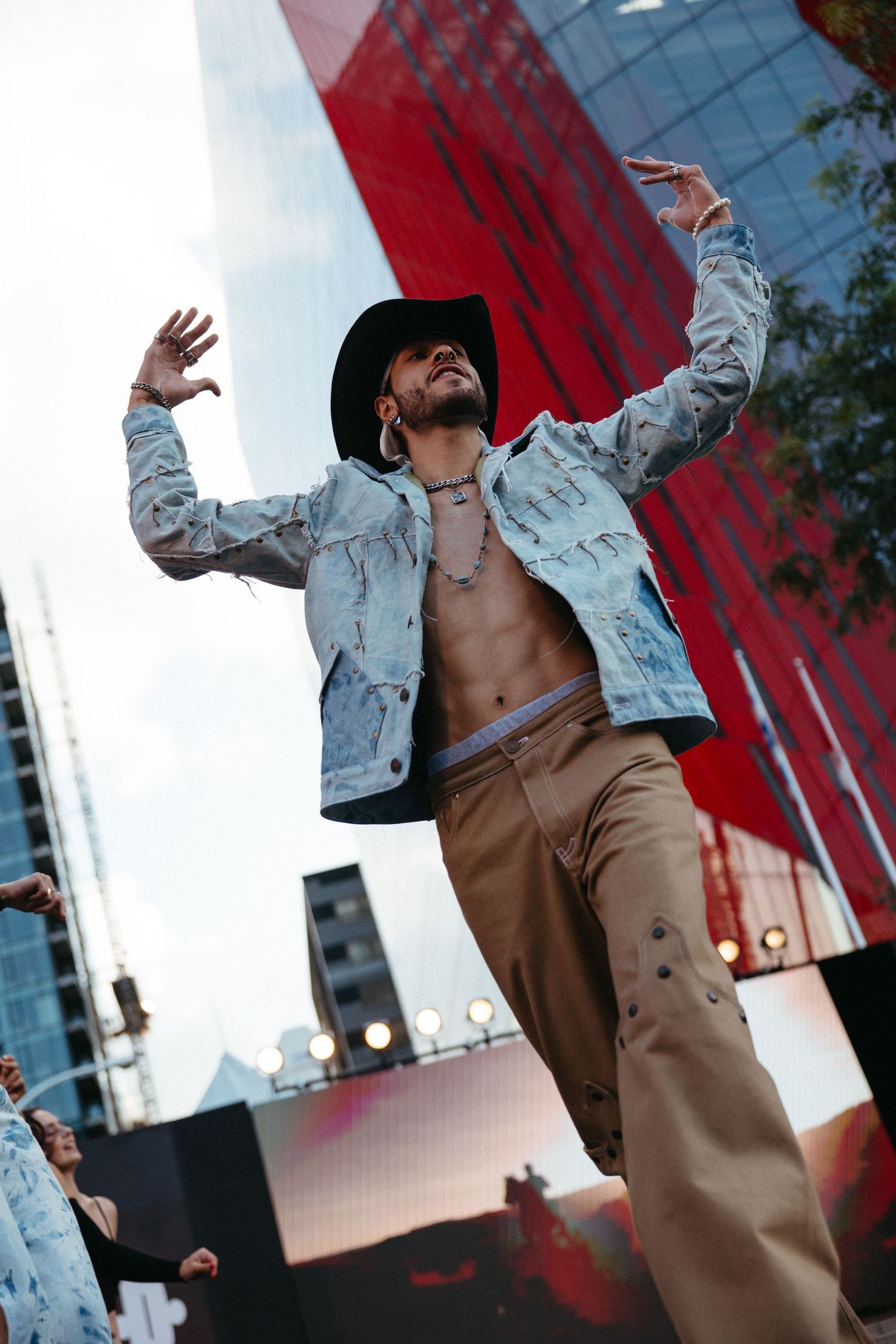 A performer in a cowboy hat and denim jacket dances energetically on stage with a red backdrop and city buildings.