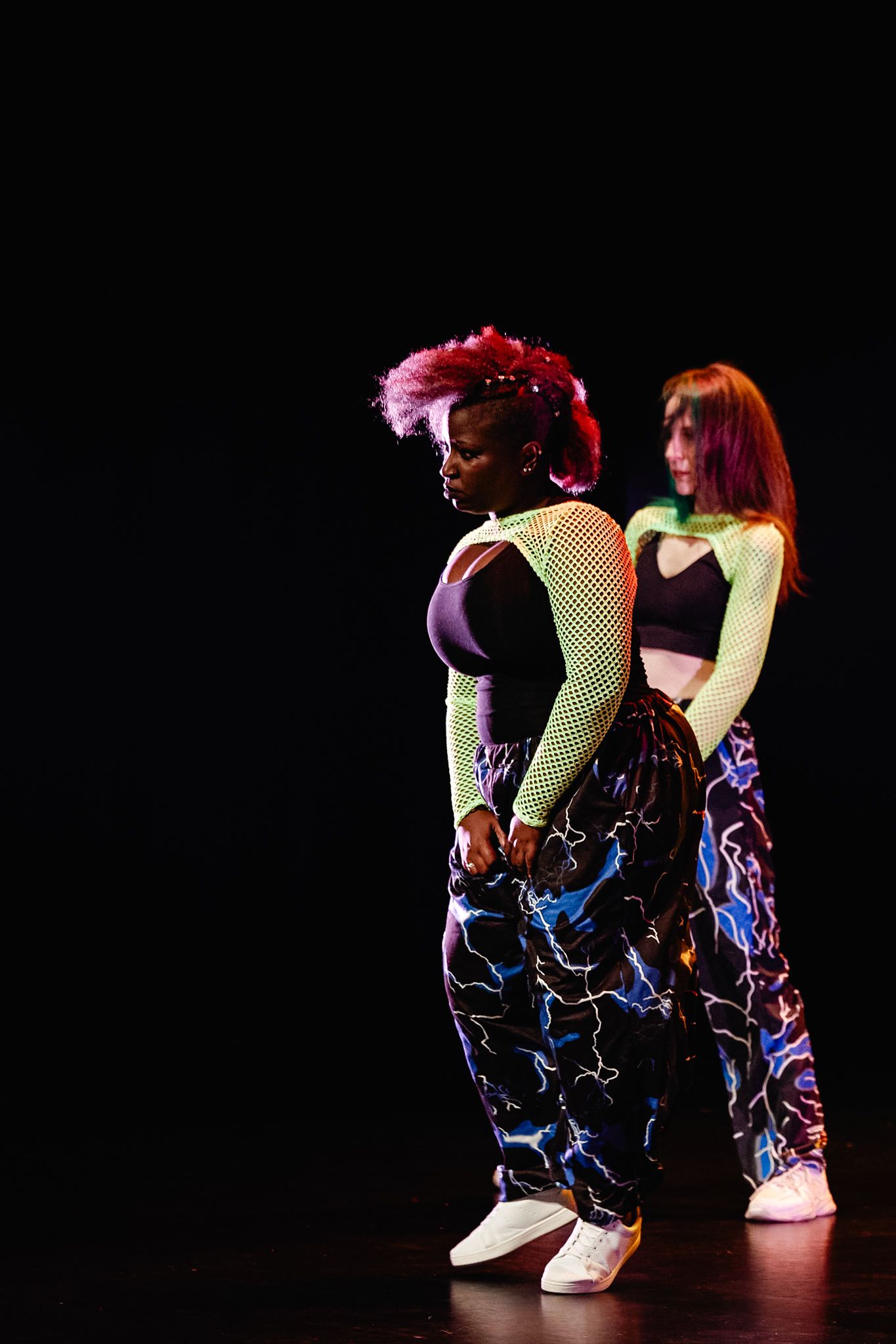 Dancers perform in sequence wearing fishnet stockings and black outfits under dramatic stage lighting.