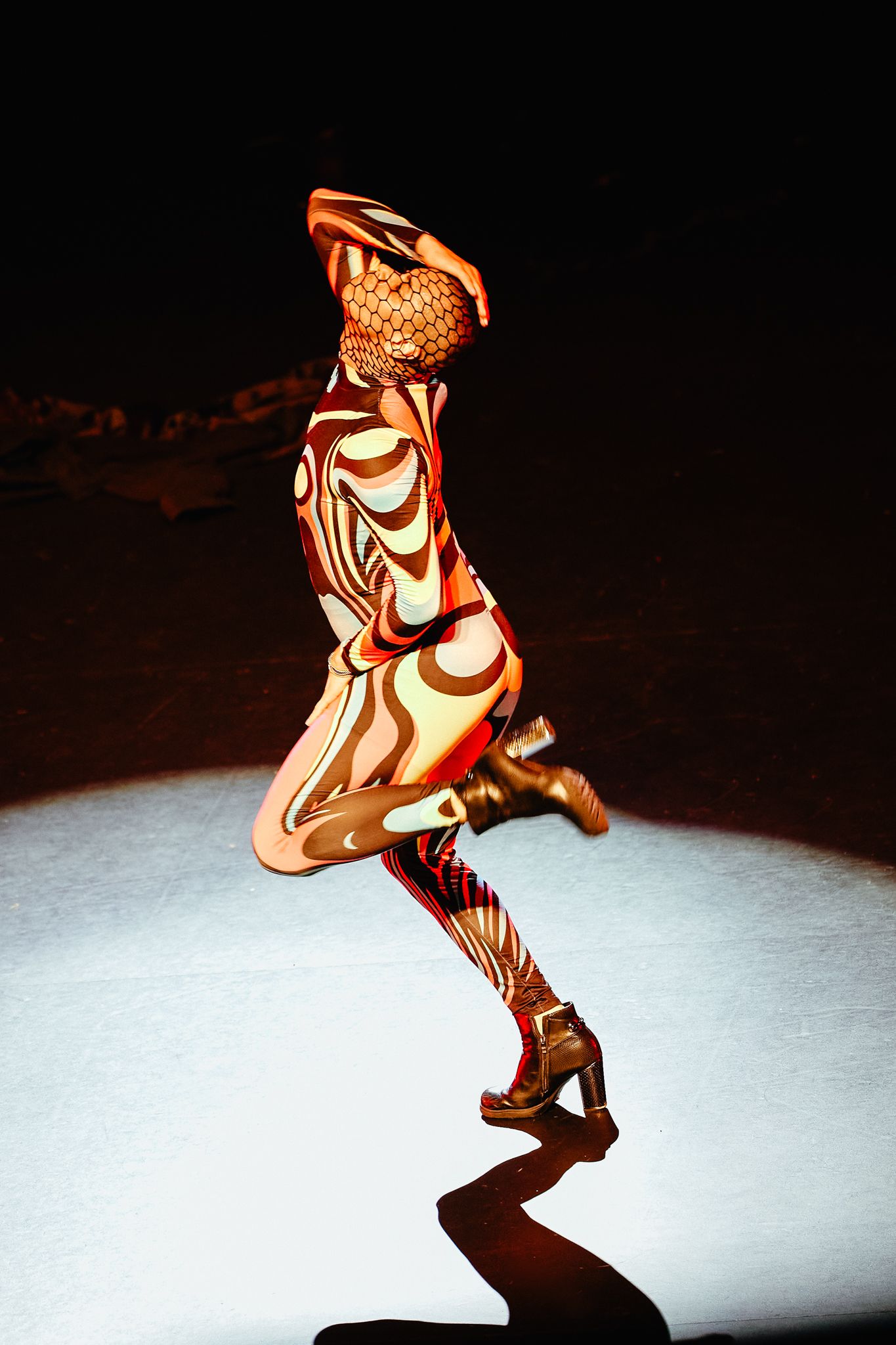 A performer in a vibrant orange and brown swirled bodysuit strikes a dramatic pose under a spotlight.