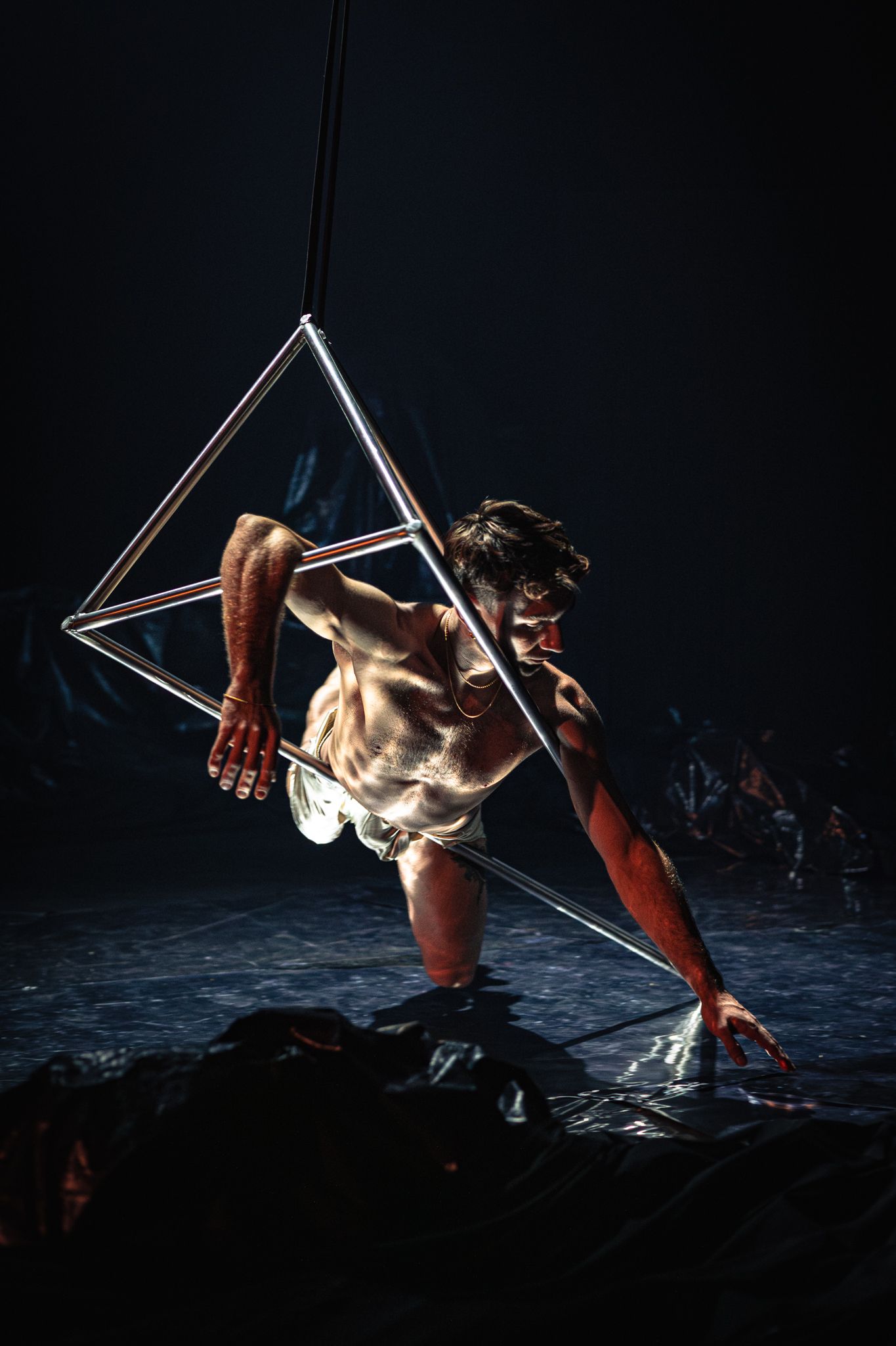Dramatic artistic performance with a performer suspended in geometric metal framework against dark background.