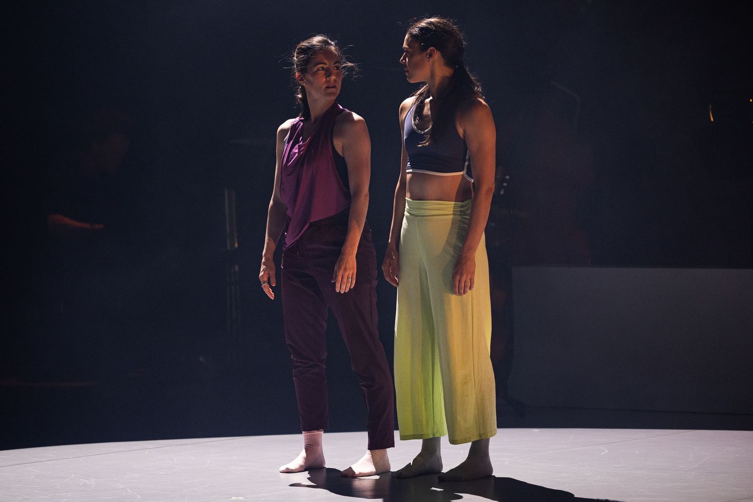 Two dancers perform in dramatic lighting on a dark stage, wearing purple and yellow flowy costumes.