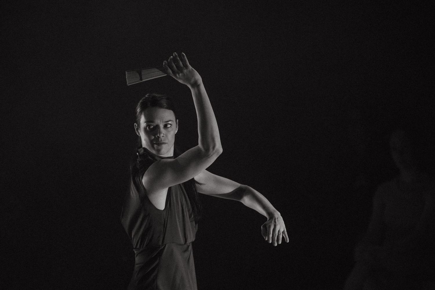 Dramatic black and white photo of a dancer in motion with raised arms against a dark background.