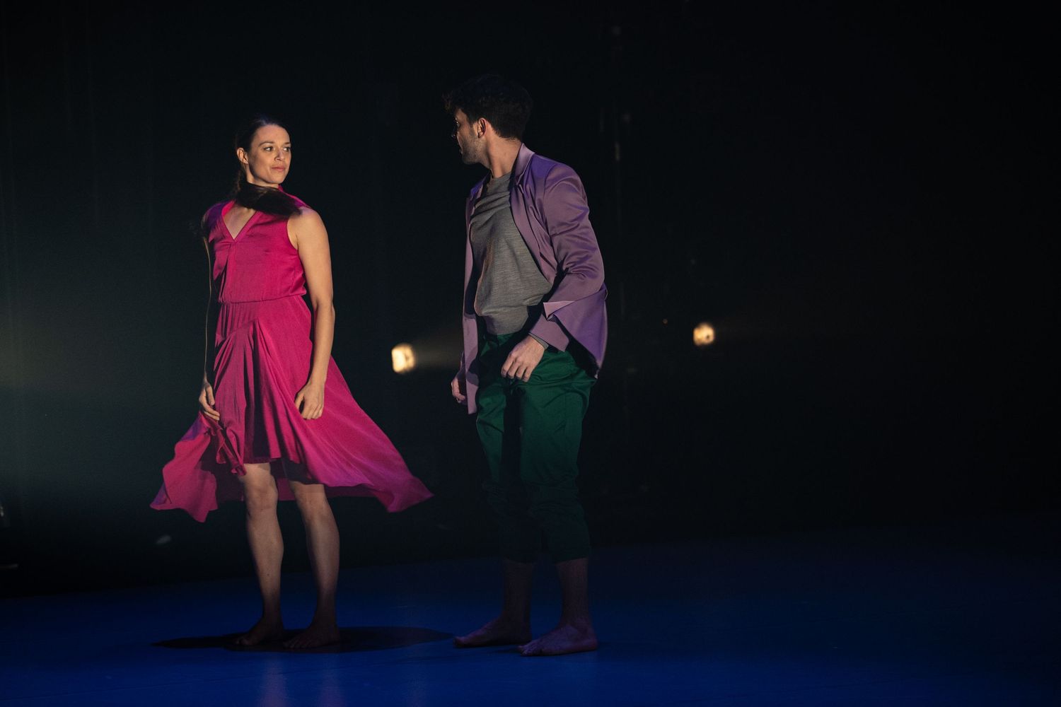 Two dancers perform on a dark stage, with one in a flowing pink dress and the other in purple attire.