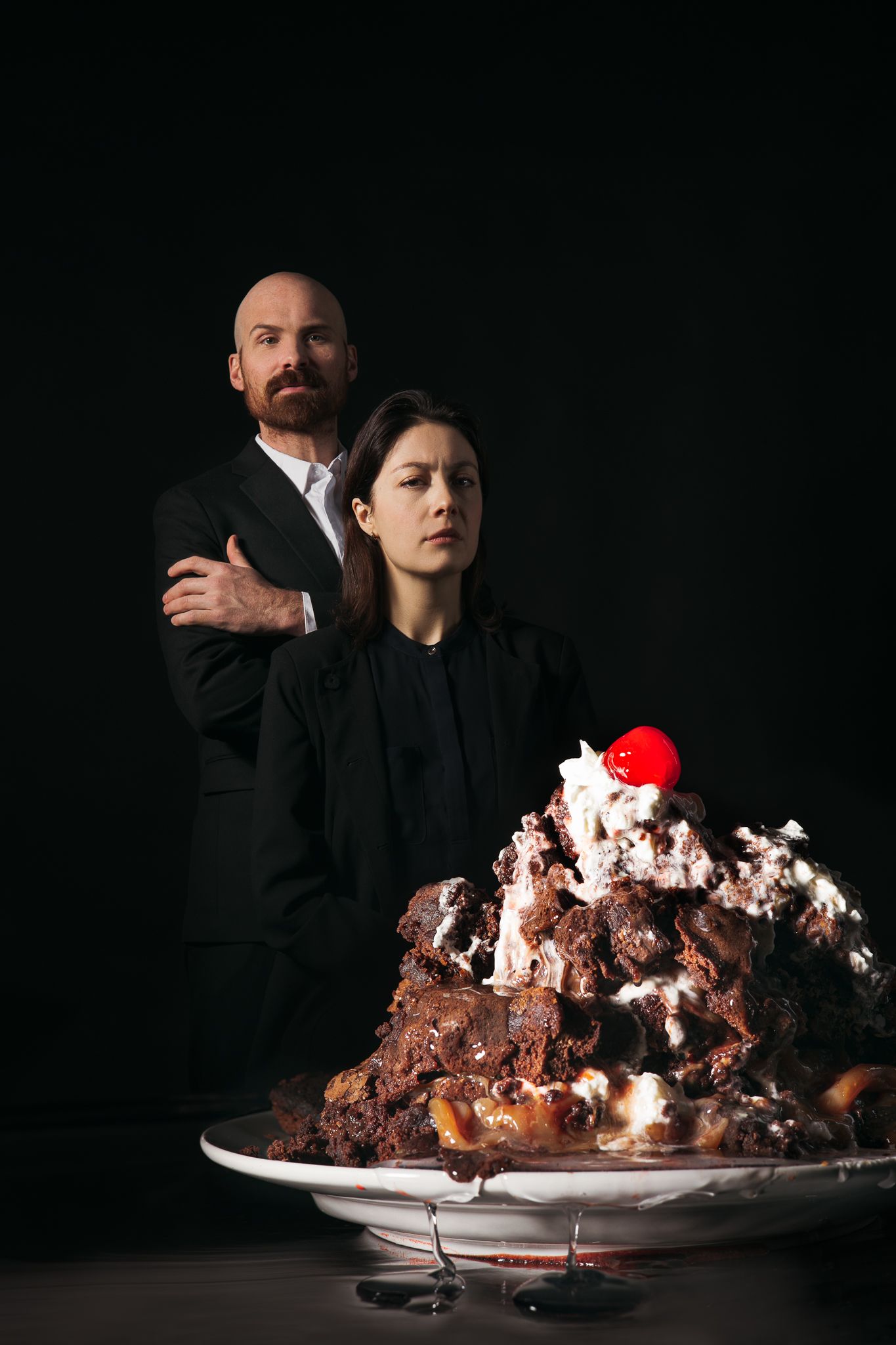 A dramatic dark portrait with a decadent chocolate dessert topped with cream and a red cherry in the foreground.