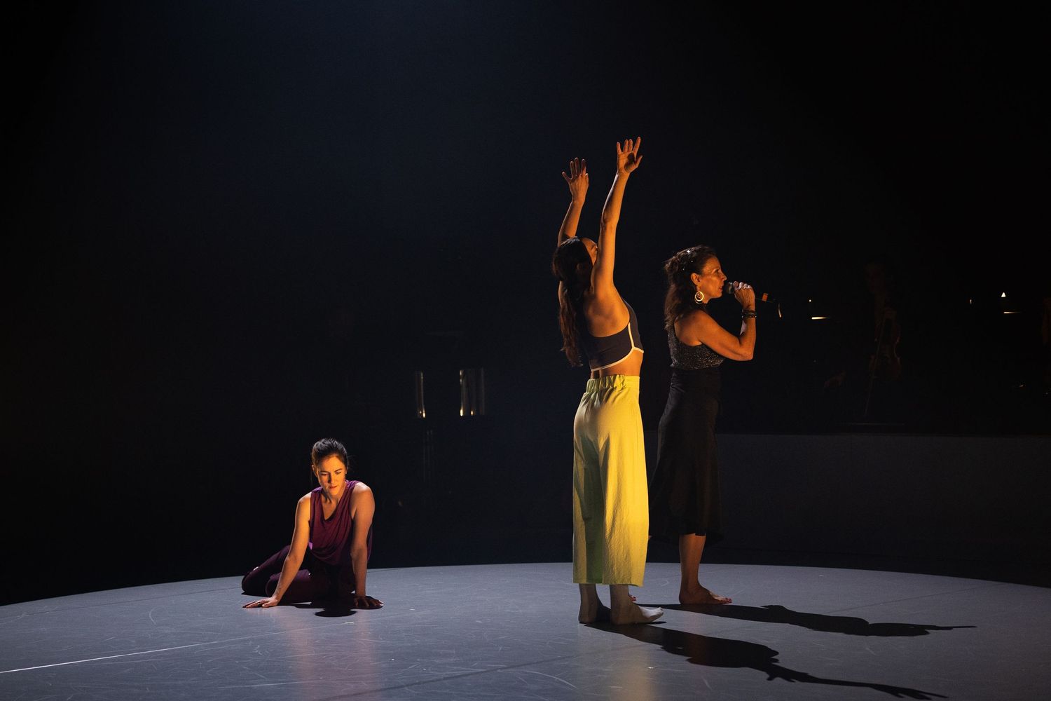 Three dancers perform on a dimly lit stage, creating dramatic shadows against a dark background.