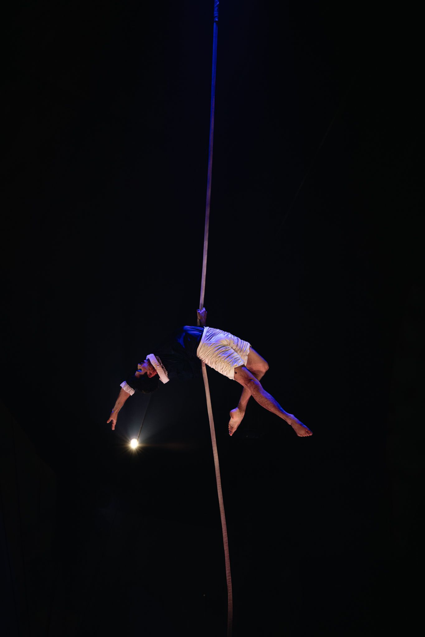 Artist performs graceful aerial acrobatics on suspended pole against dark background.