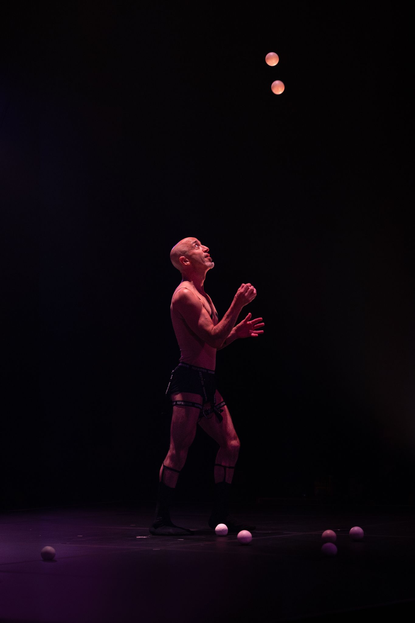 A performer juggles balls under dramatic red stage lighting against a dark background during a theatrical performance.