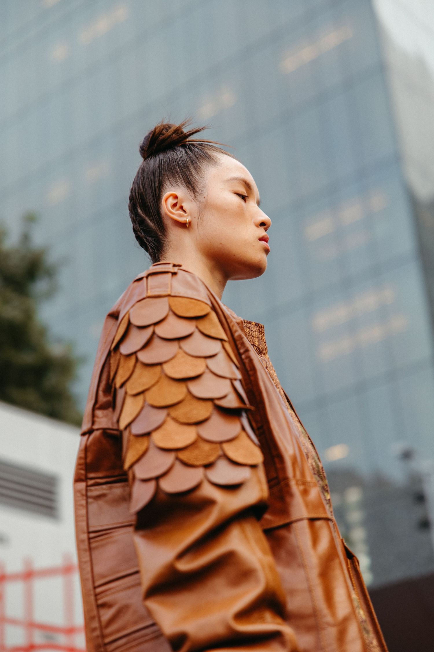 Fashion model in brown leather jacket with scale-like pattern detail poses against urban skyscraper background.