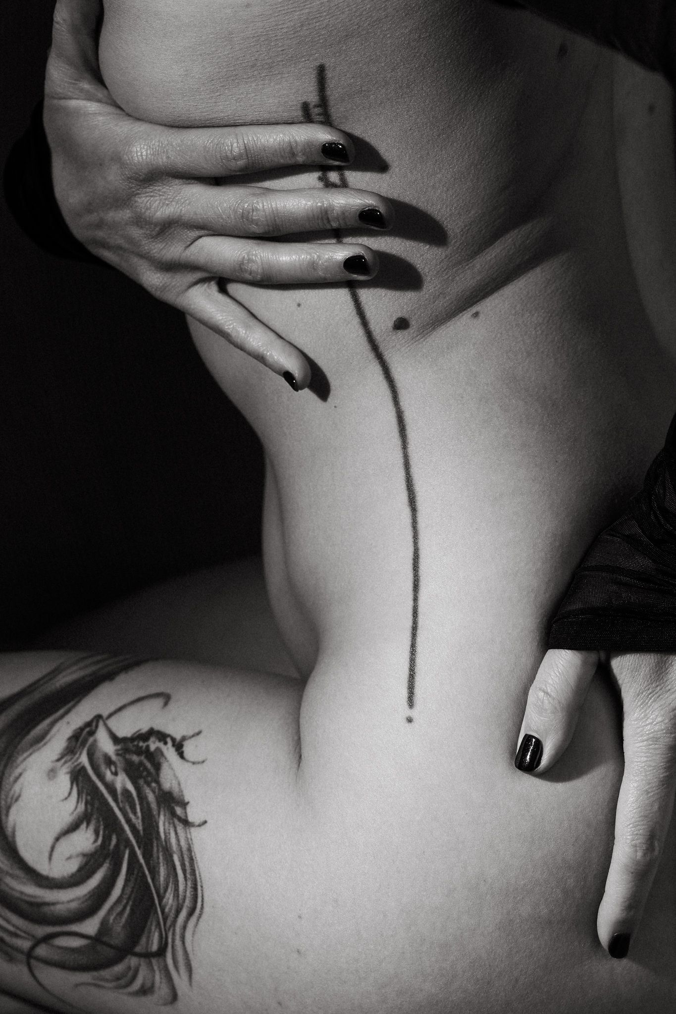 Black and white artistic photo showing hands with dark nails and delicate linework tattoo details.